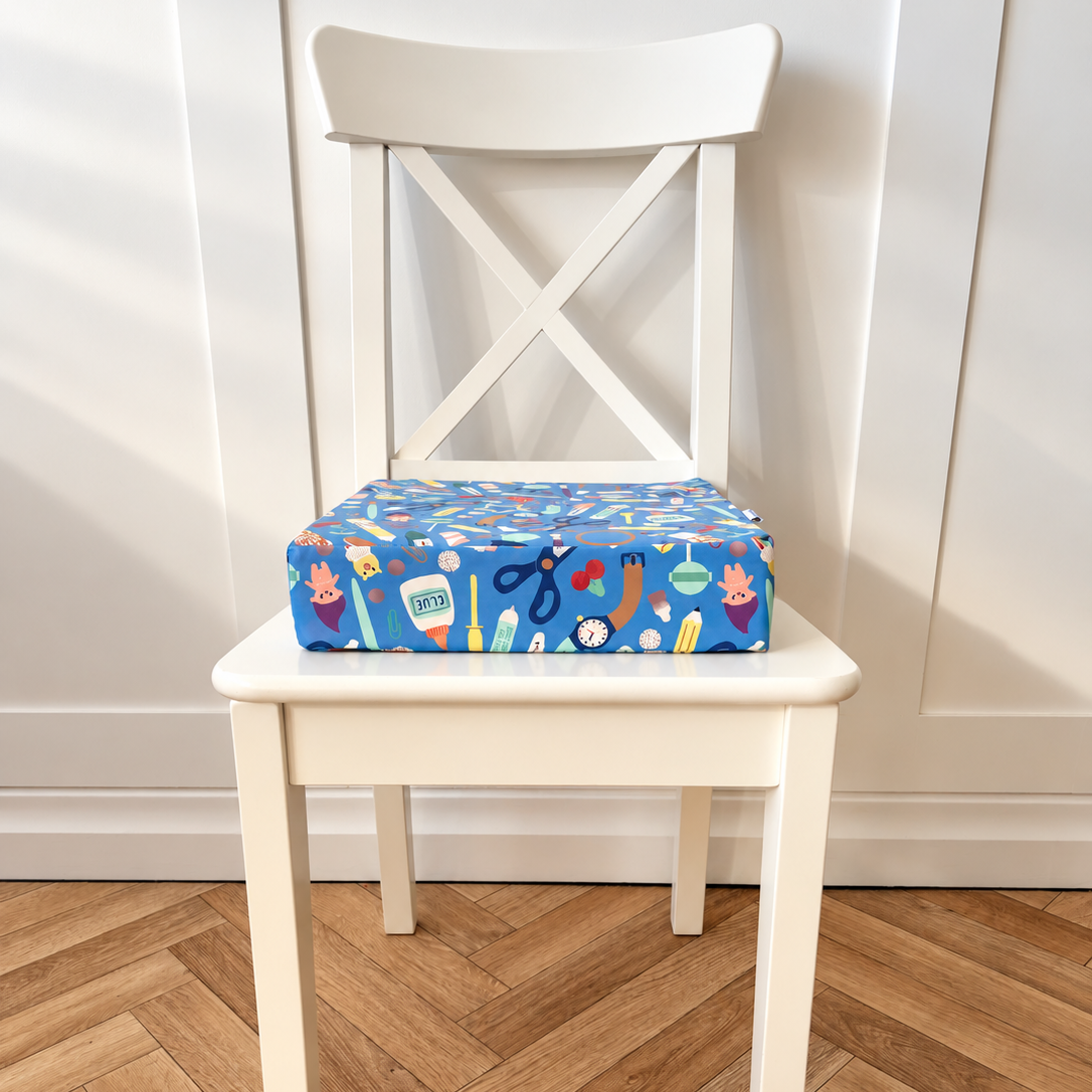 A square blue booster cushion with a colourful craft-themed pattern sits on a white wooden chair in front of a white panelled wall and herringbone wood floor.