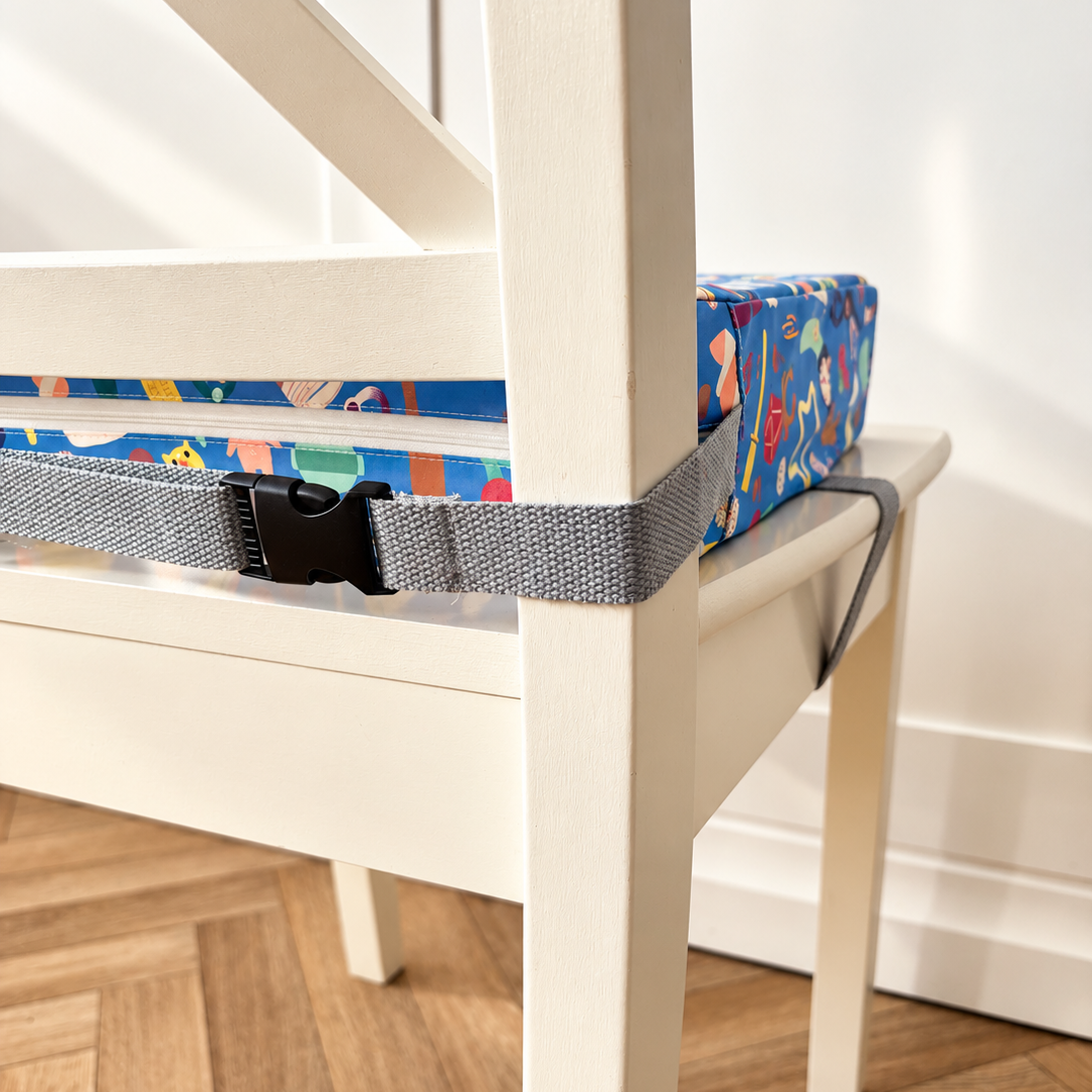 A close-up of a white wooden chair with a grey adjustable seat strap securing a colourful patterned Uppy booster cushion in place.