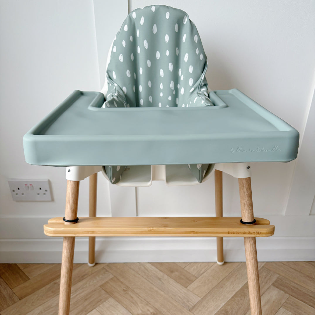 Children's high chair with a green seat and wooden legs on a wooden floor.