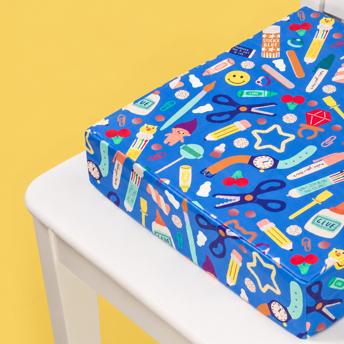 A blue booster cushion with a colourful school-themed pattern, displayed on a white chair against a solid yellow background.