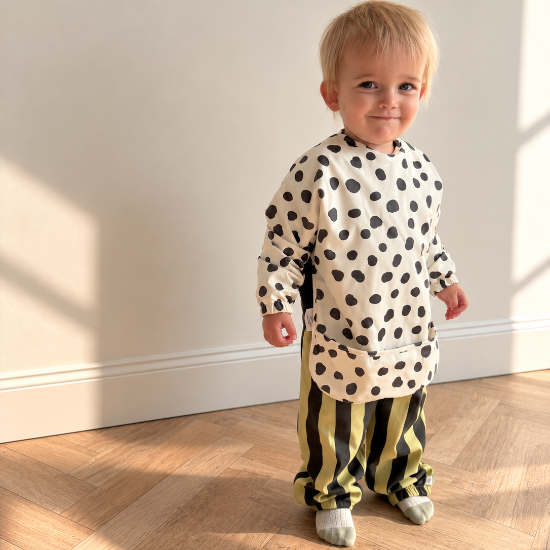 A toddler standing indoors on a light wood floor against a plain wall, wearing a cream long-sleeve bib with black polka dots and black-and-green vertically striped pants.