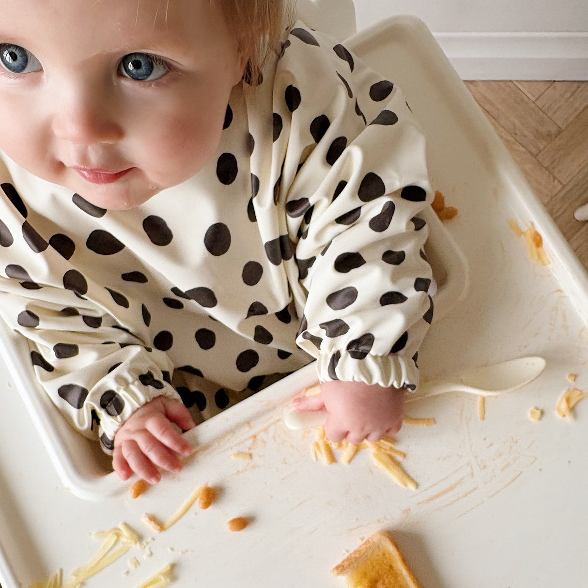 Baby in a highchair looking up. Food on the tray and wearing a white and black spotty bib