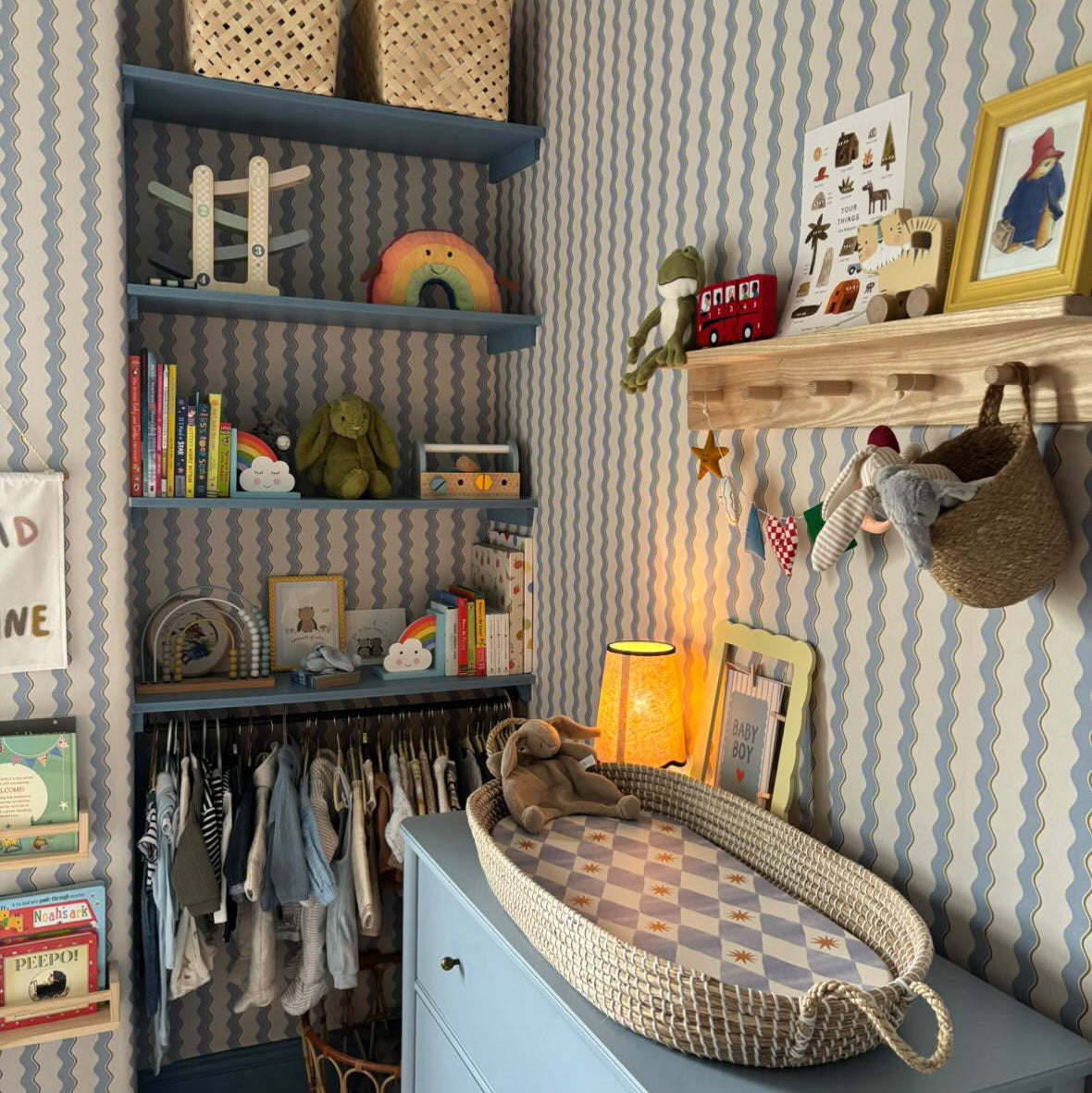 Nursery room changing area with blue squiggle wallpaper, blue shelfs and baby clothes hung up