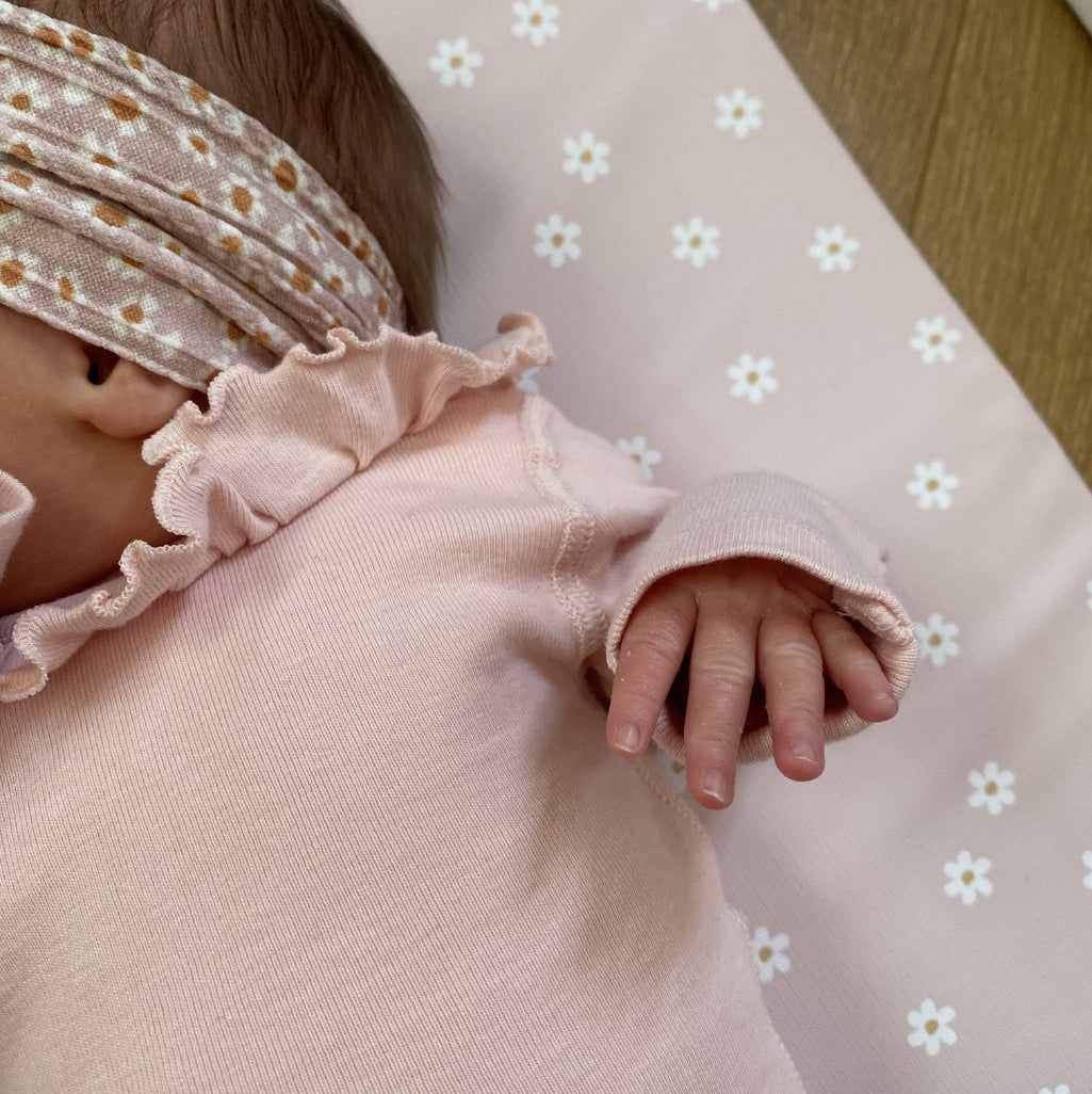Baby wearing a pink outfit with ruffled details on a light-colored pink changing mat with daisy floral pattern.