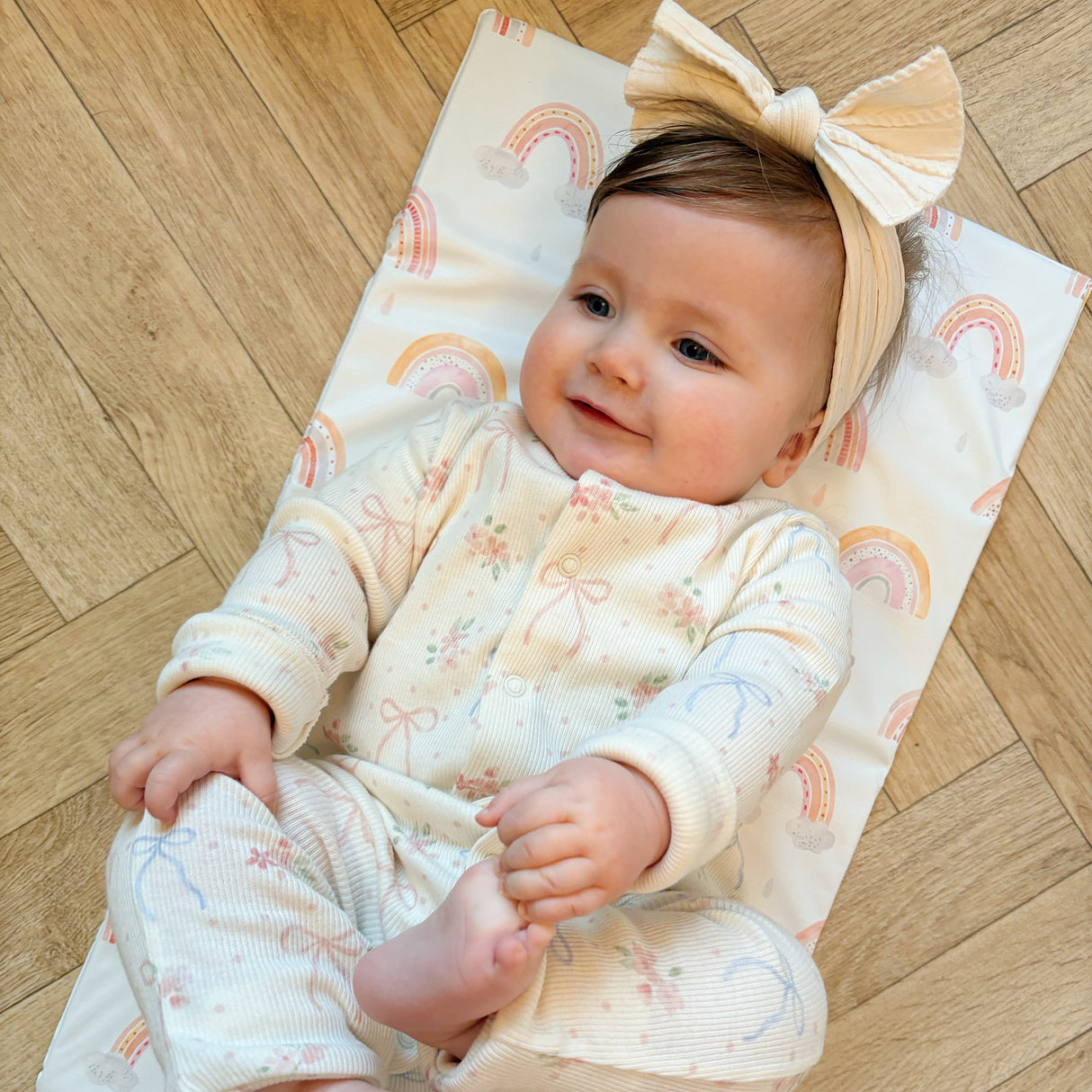 Baby wearing a bow headband laying on a changing mat with rainbow pattern on a wooden floor.