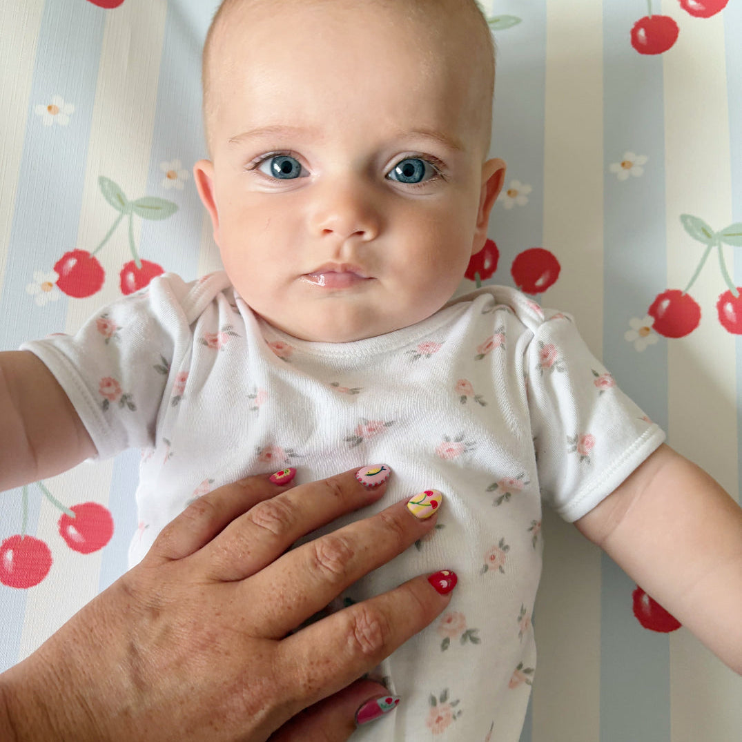 Baby in a white floral shirt with a hand on its chest, standing against a striped background with red cherries.