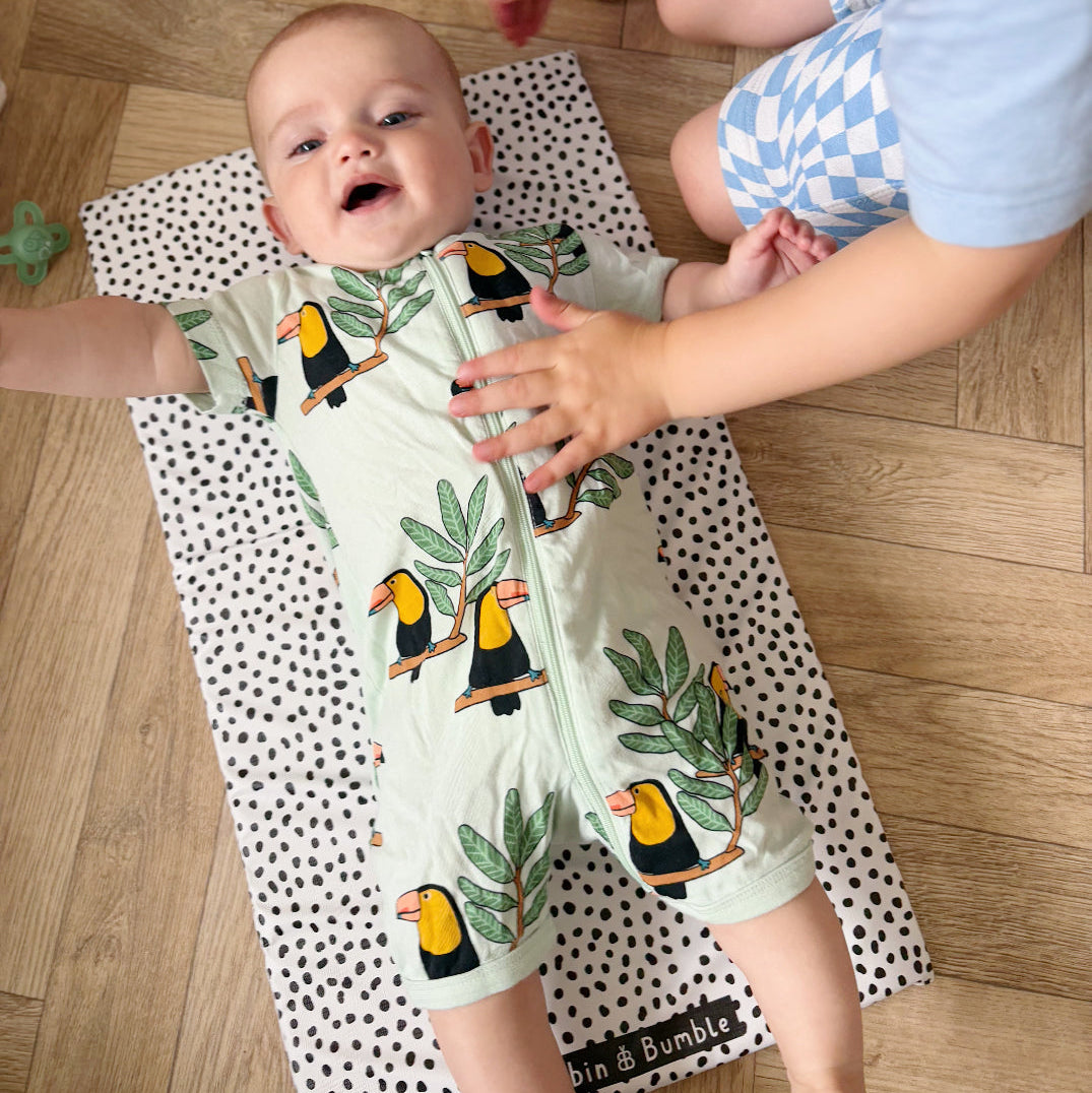 Baby wearing a onesie with toucan design laying on a polka dot changing mat on a wooden floor