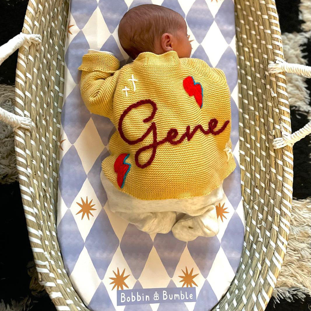Baby in a yellow knitted outfit with 'Gene' embroidered in a woven basket on a patterned fabric background.
