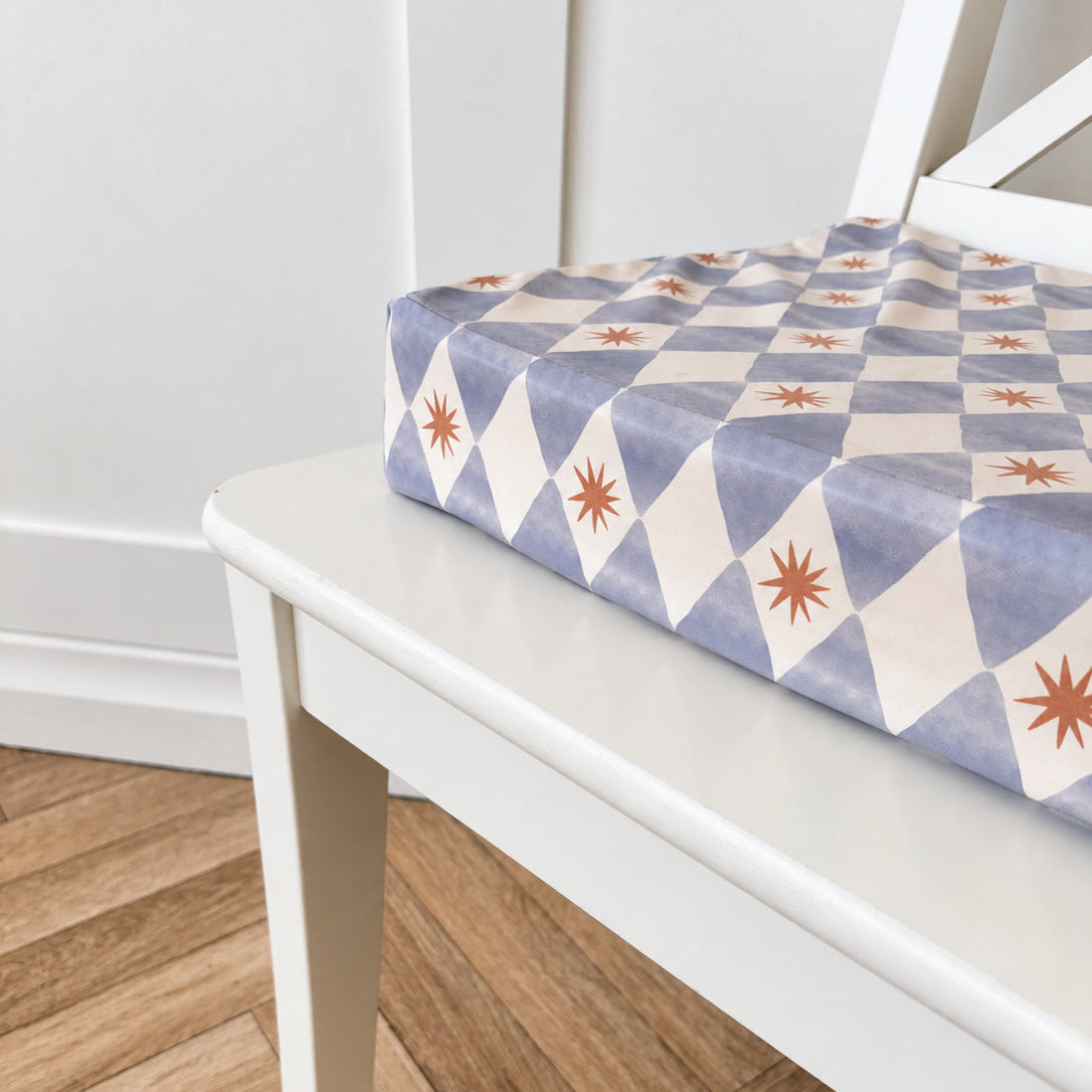A close-up of a patterned fabric booster cushion or pad resting on a white wooden chair in a room with a light wood herringbone floor.