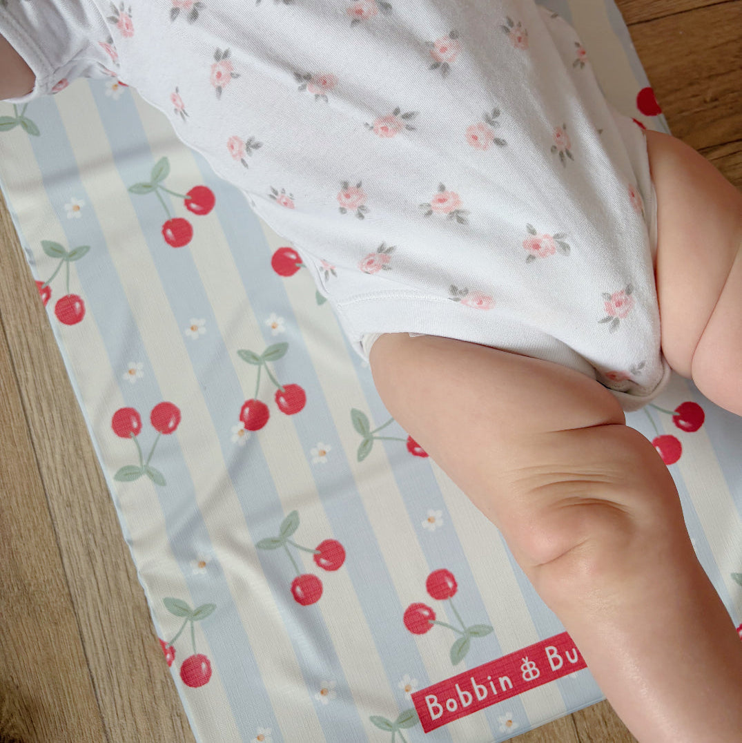 Baby in a floral outfit on a cherry-patterned changing mat with 'Bobbin & Bumble' branding.