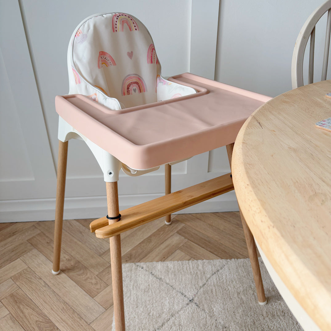 Pink high chair with a white cushion featuring rainbow patterns, placed next to a wooden table on a wooden floor.