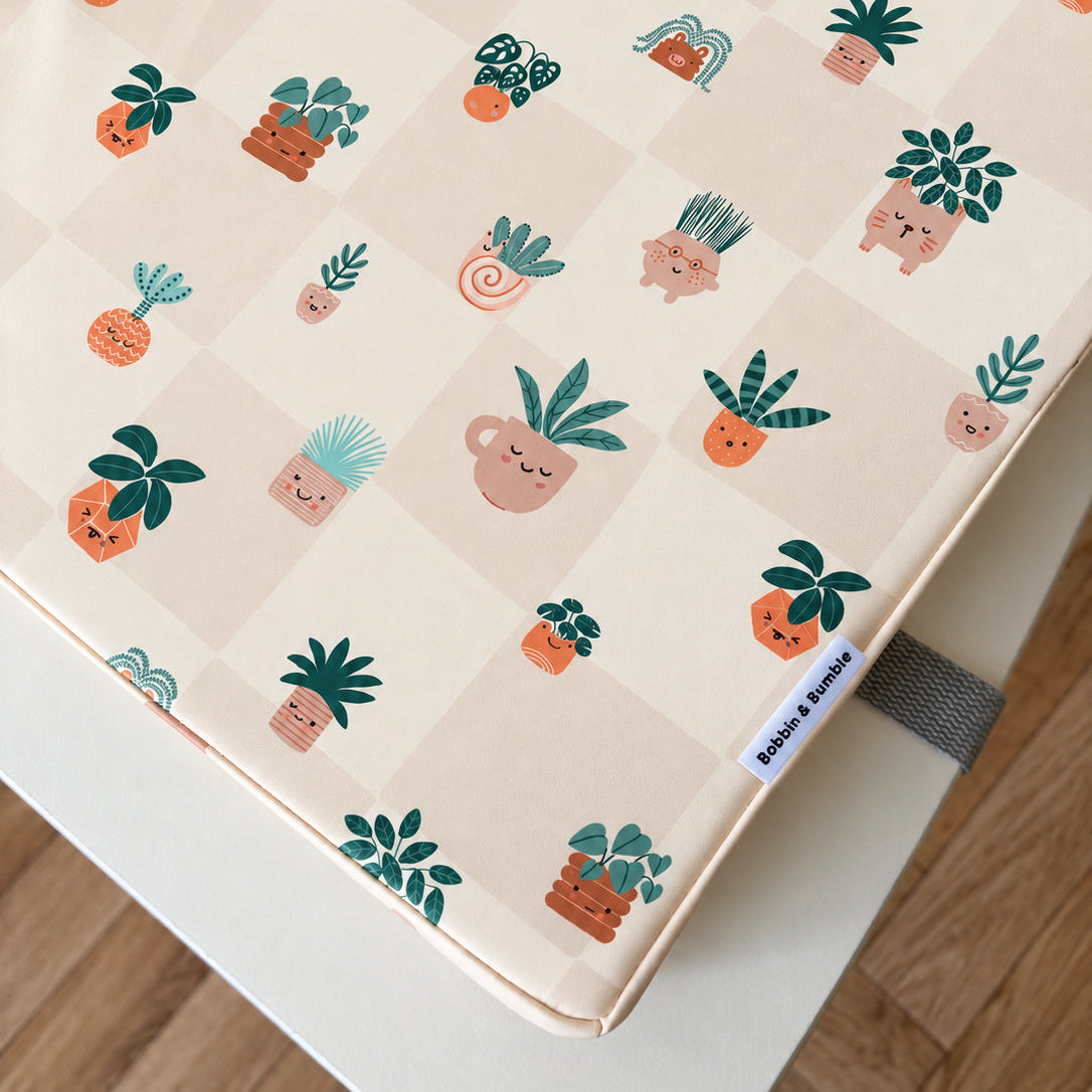 A close-up of a toddler booster pad with a cute, repeating pattern of smiling potted plants on a cream-colored checkered background. The brand name 'Bobbin and Bumble' is visible on a small fabric tag on the right side.