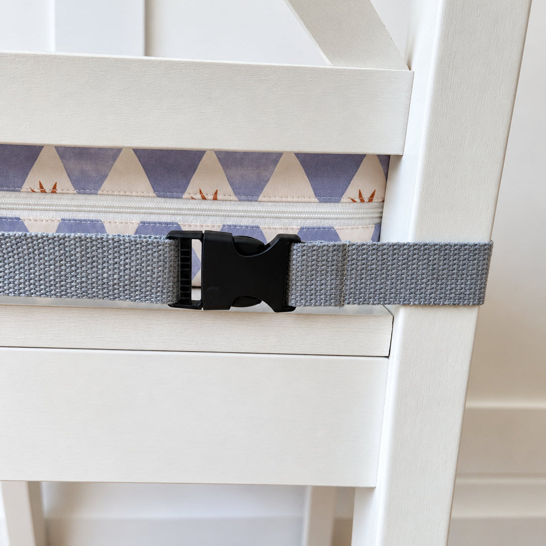 A gray webbed safety restraint strap with a black plastic buckle, shown attached to a white wooden chair in a home setting with a wood floor visible.