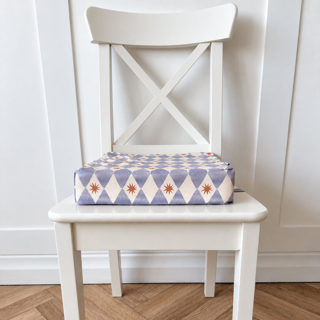 A white wooden chair with a patterned booster cushion on the seat. The cushion features a blue-and-white harlequin diamond pattern with small mustard yellow starburst accents.