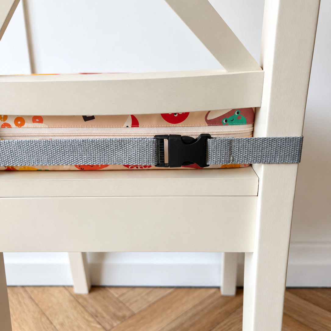 A close-up of a gray adjustable seat belt with a black buckle strapped across the seat of a cream-colored wooden chair. The chair is set against a white wall and light wood herringbone floor.