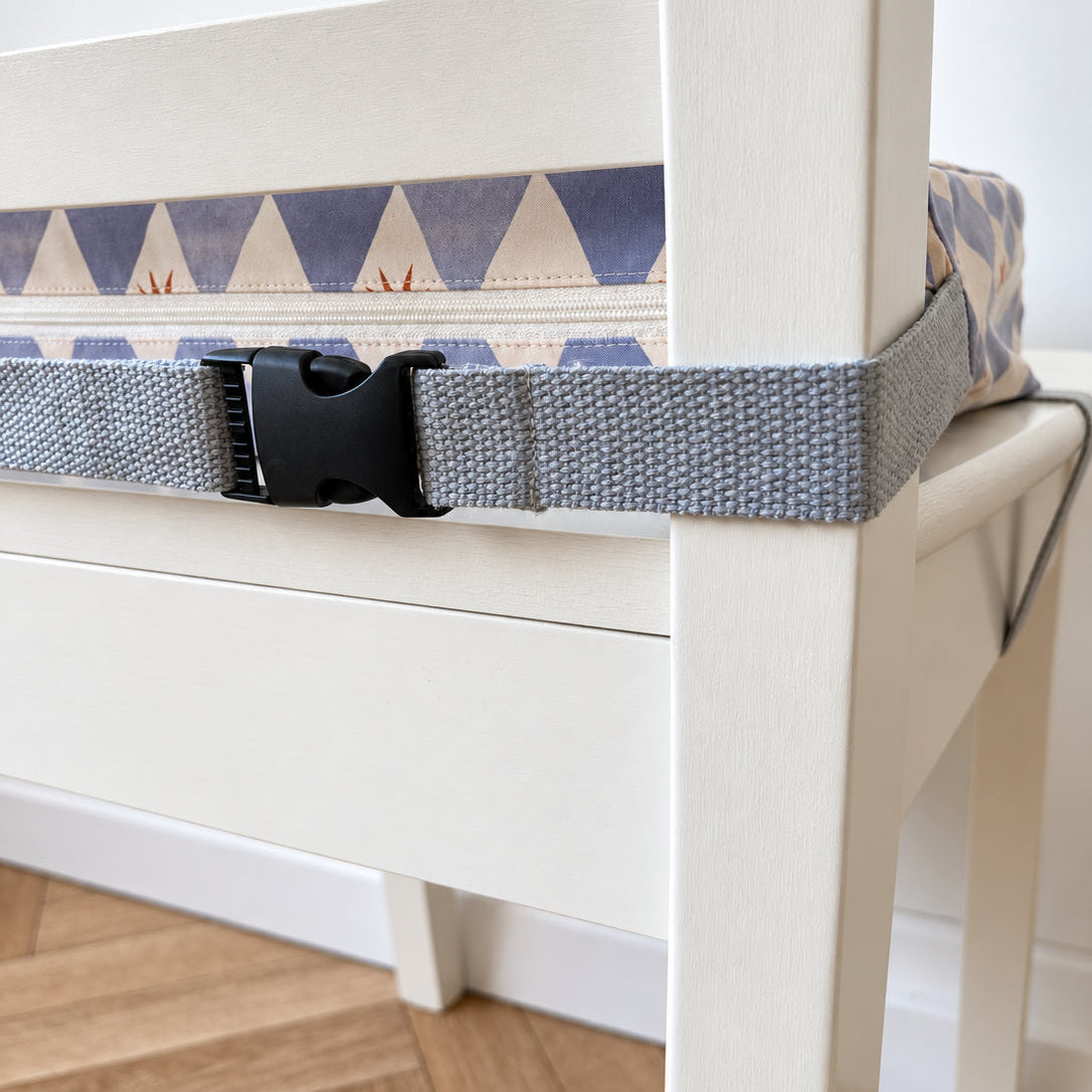 A close-up of a grey woven seat belt with a black plastic buckle strapped across the seat of a white wooden chair, with a patterned booster cushion visible underneath.