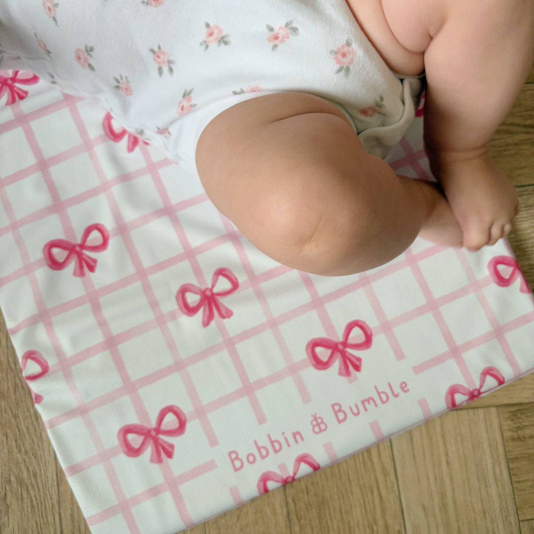 Baby's legs on a pink and white checkered changing mat with bow pattern, featuring 'Bobbin & Bumble' branding.