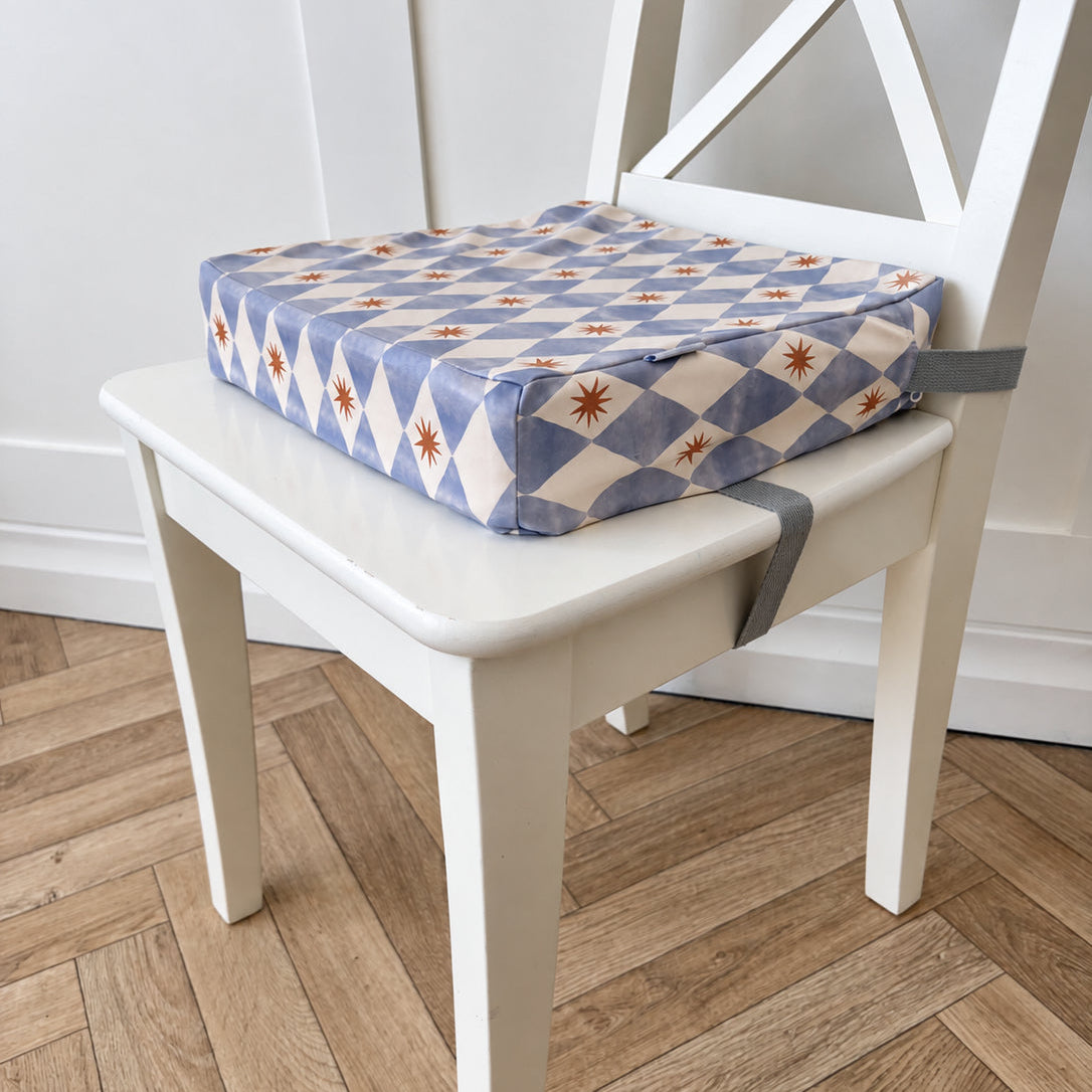 A square toddler booster cushion with a blue, cream, and yellow diamond pattern sits on a white wooden chair in a room with a light wood herringbone floor.