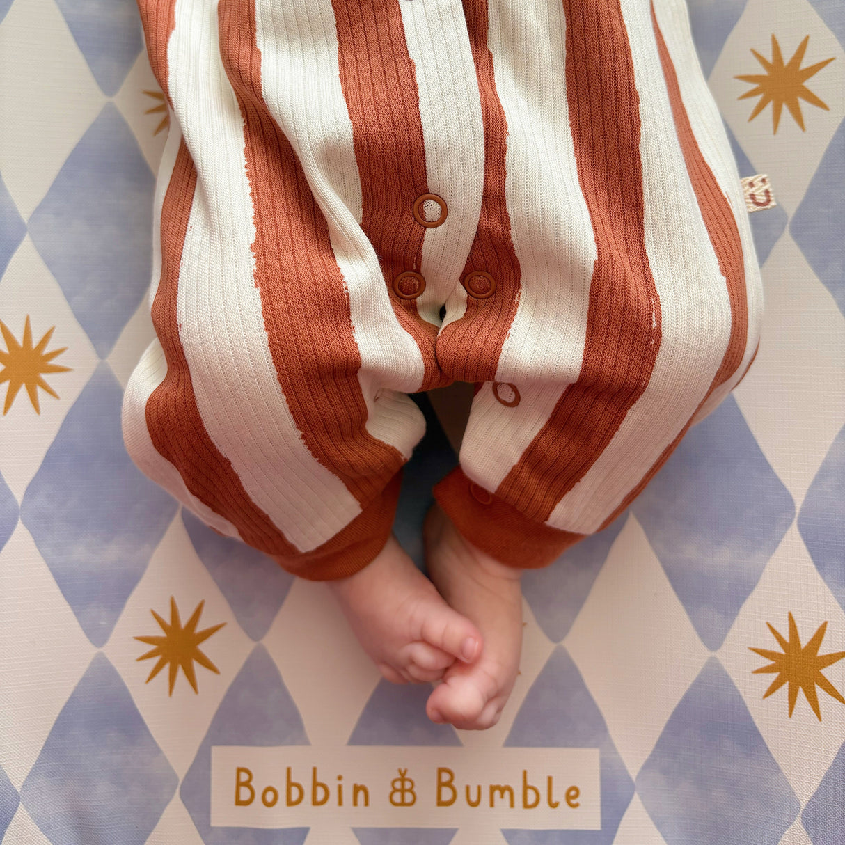 Baby wearing brown striped sleepsuit, on a blue harlequin changing mat with Bobbin and Bumble branding.