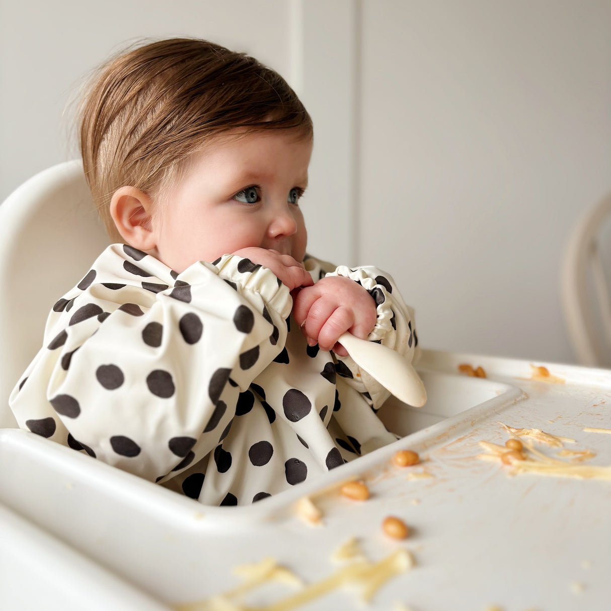 Baby in a highchair wearing a spotty long sleeved bib with food on the highchair tray