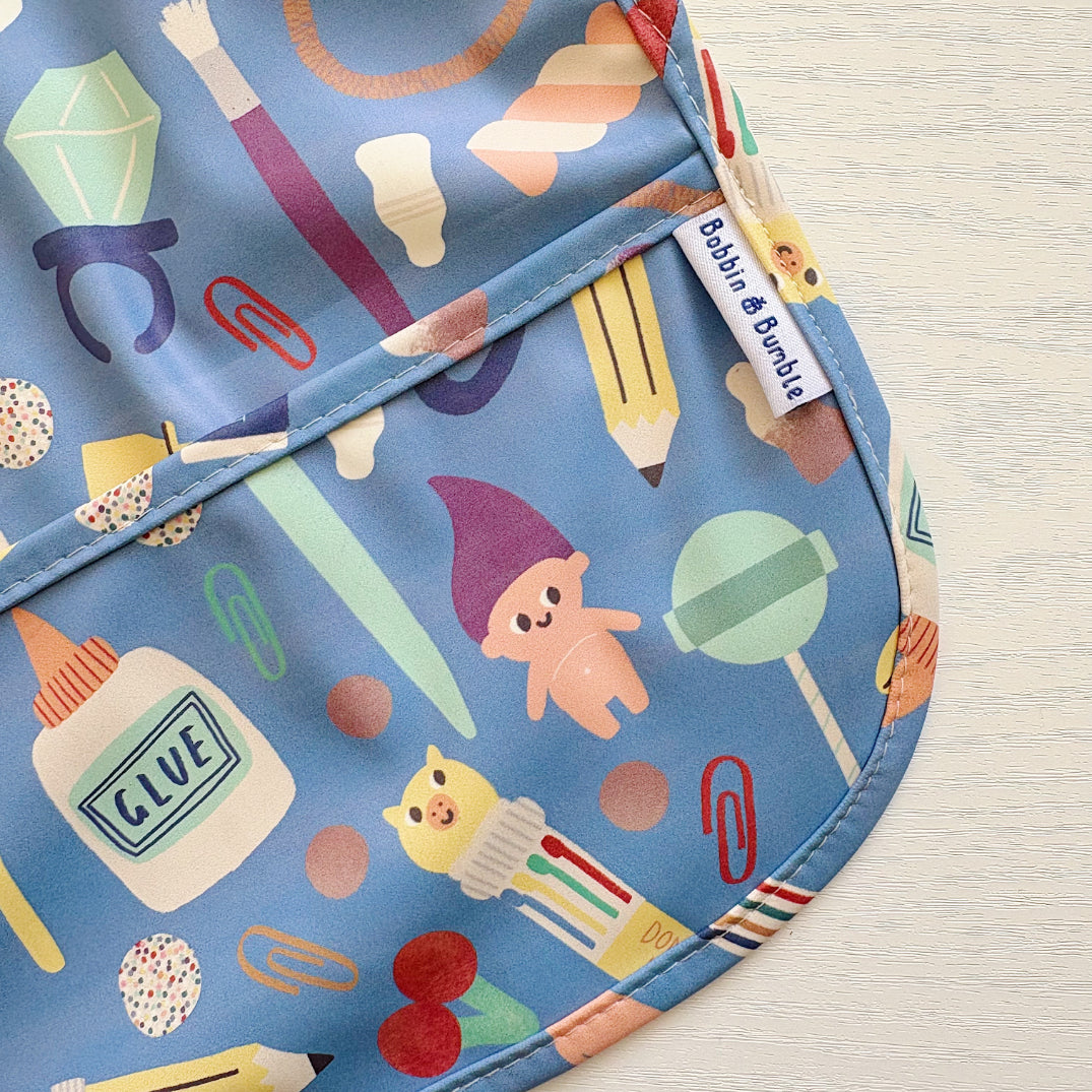 Children's apron with colourful stationery-themed design on a light wooden surface. Bobbin and Bumble Label