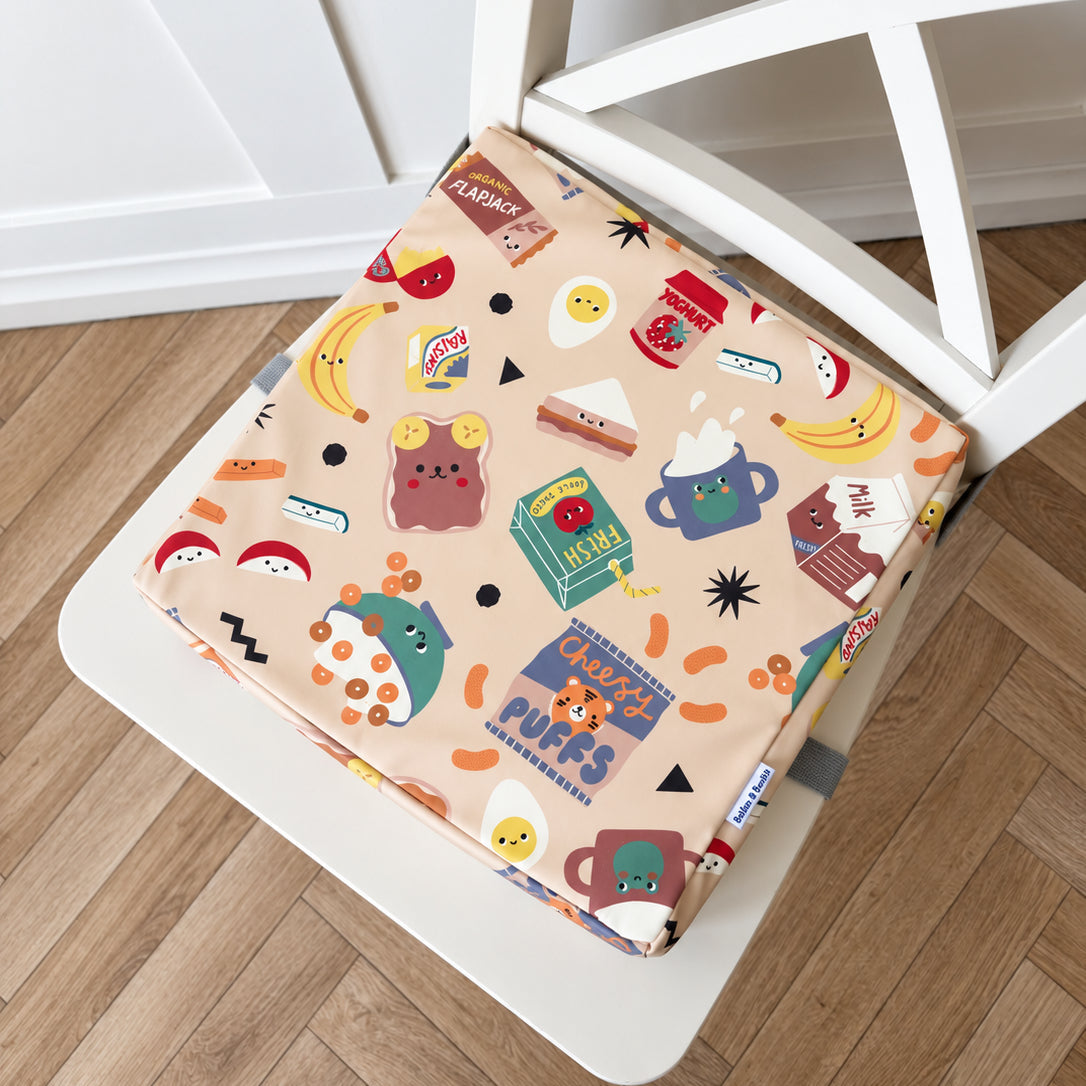 A square fabric chair booster cushion with a colourful cartoon food and drink pattern, shown resting on a white wooden chair over a light wood floor.