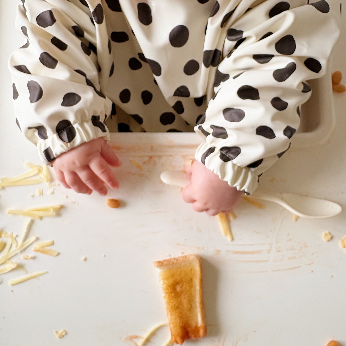 Close up of baby grabbing a spoon in a highchair with food. Wearing a long sleeved spotted bib
