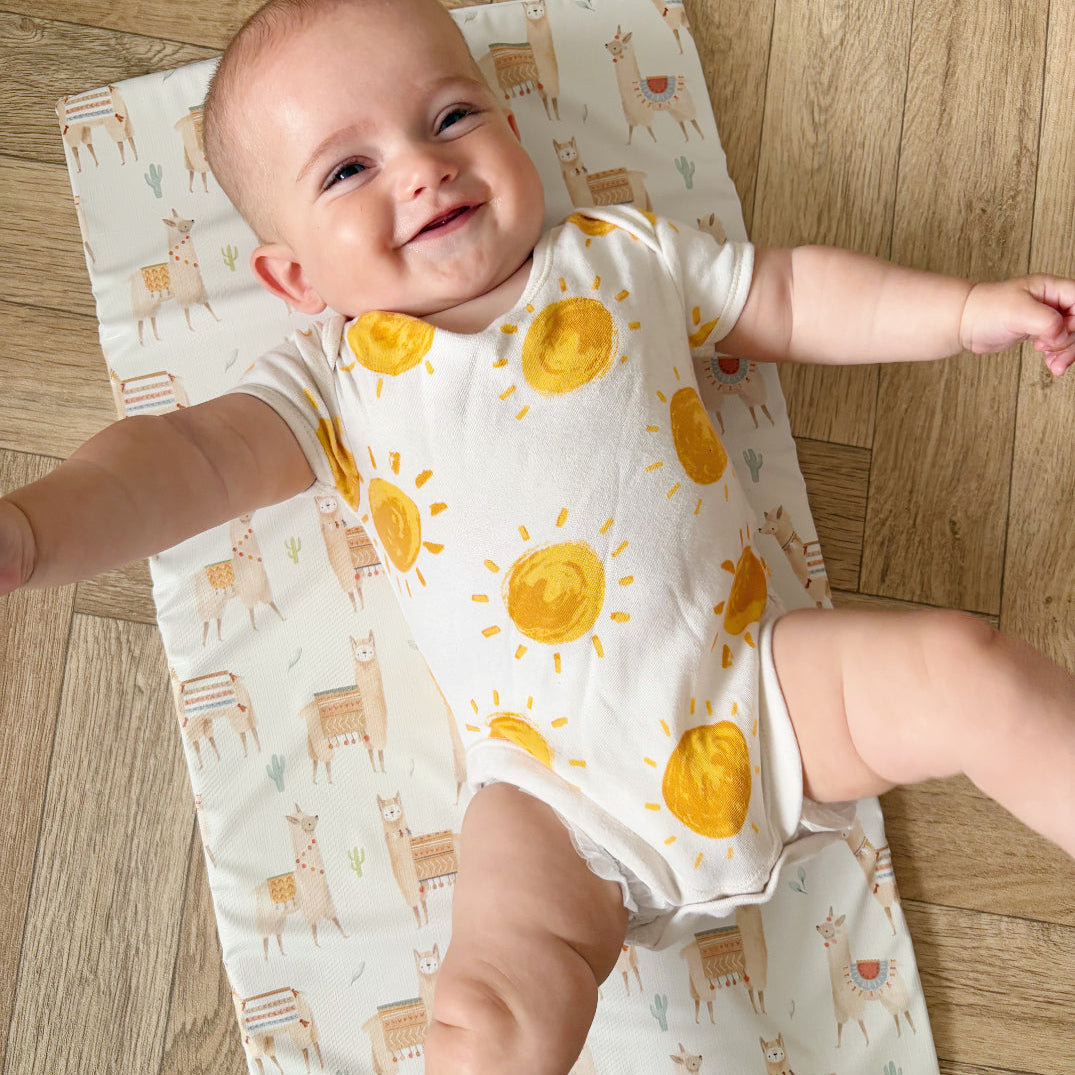 Baby in a sun-patterned onesie on a travel changing mat with alpaca designs on a wooden floor.