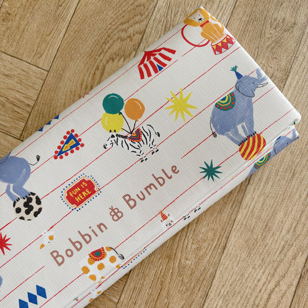 Folded changing mat with colourful animal circus patterns and 'Bobbin & Bumble' branding on a wooden floor.