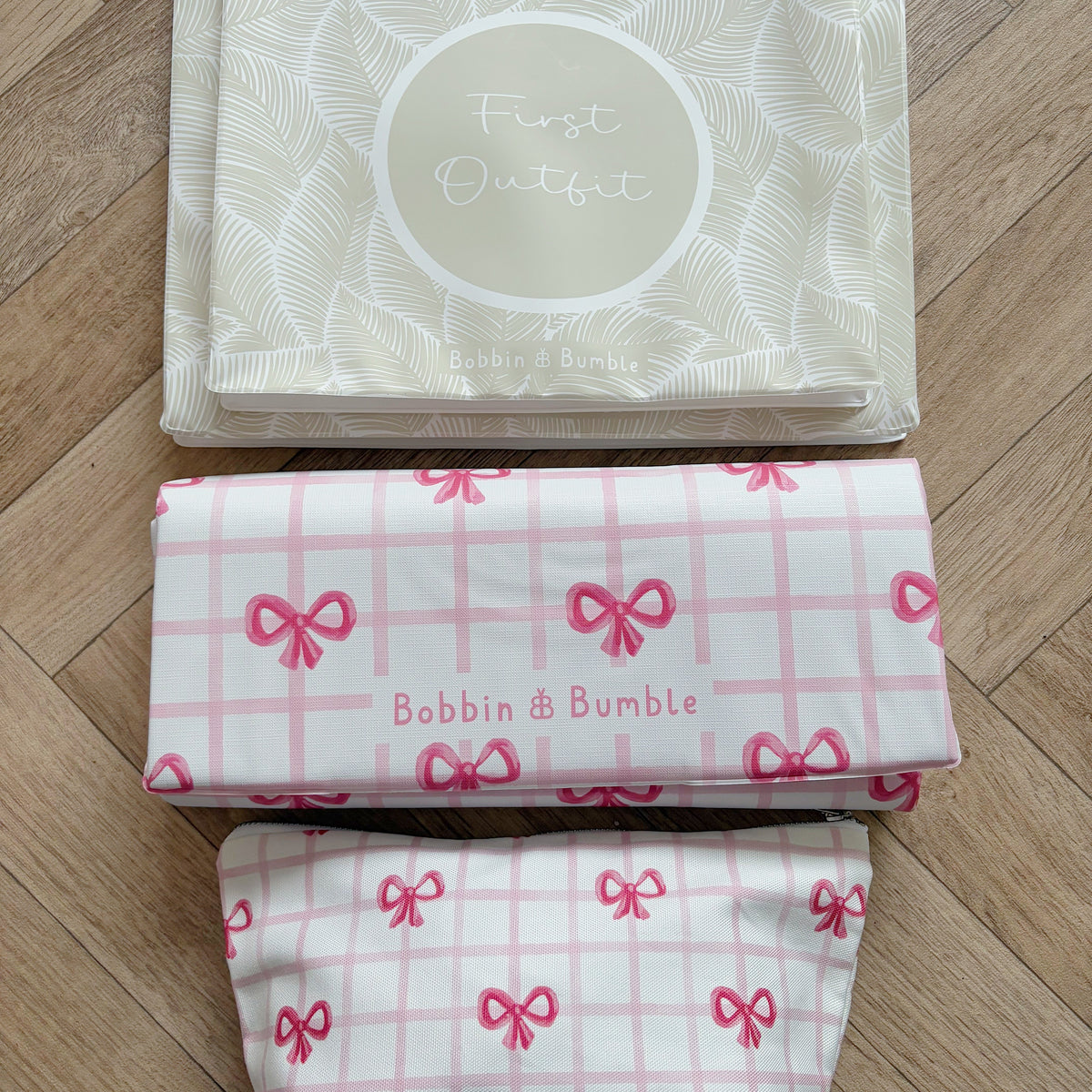 Set of hospital bags with 'Bobbin & Bumble' branding, folded travel baby changing mat and matching nappy wallet in pink bow pattern on a wooden surface