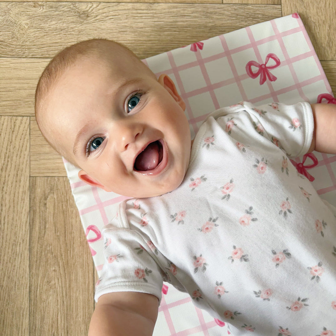 Baby lying on a pink and white checkered changing mat with red bows on a wooden floor.