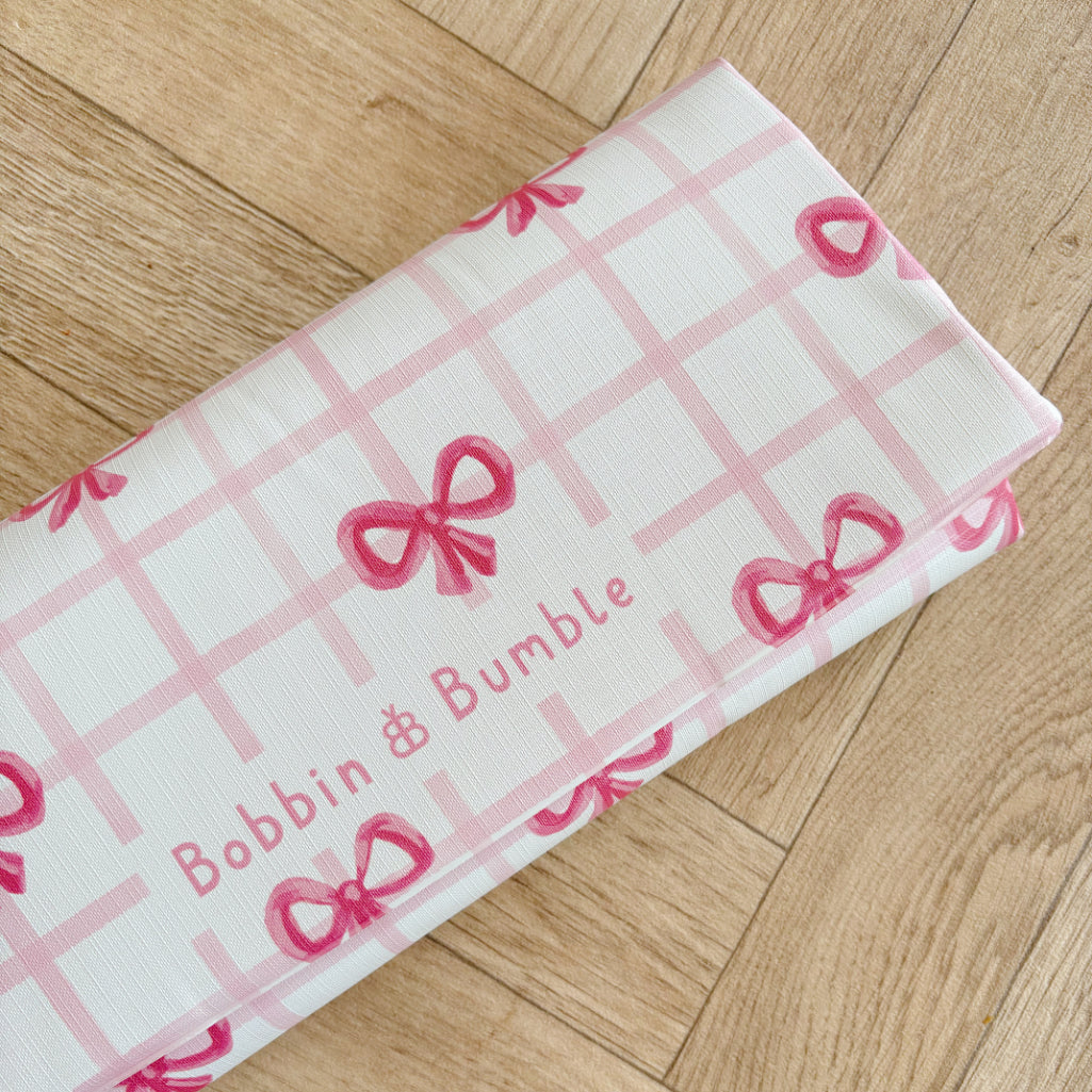 Travel changing mat with pink checkered pattern and bow designs on a wooden surface, featuring 'Bobbin & Bumble' branding.