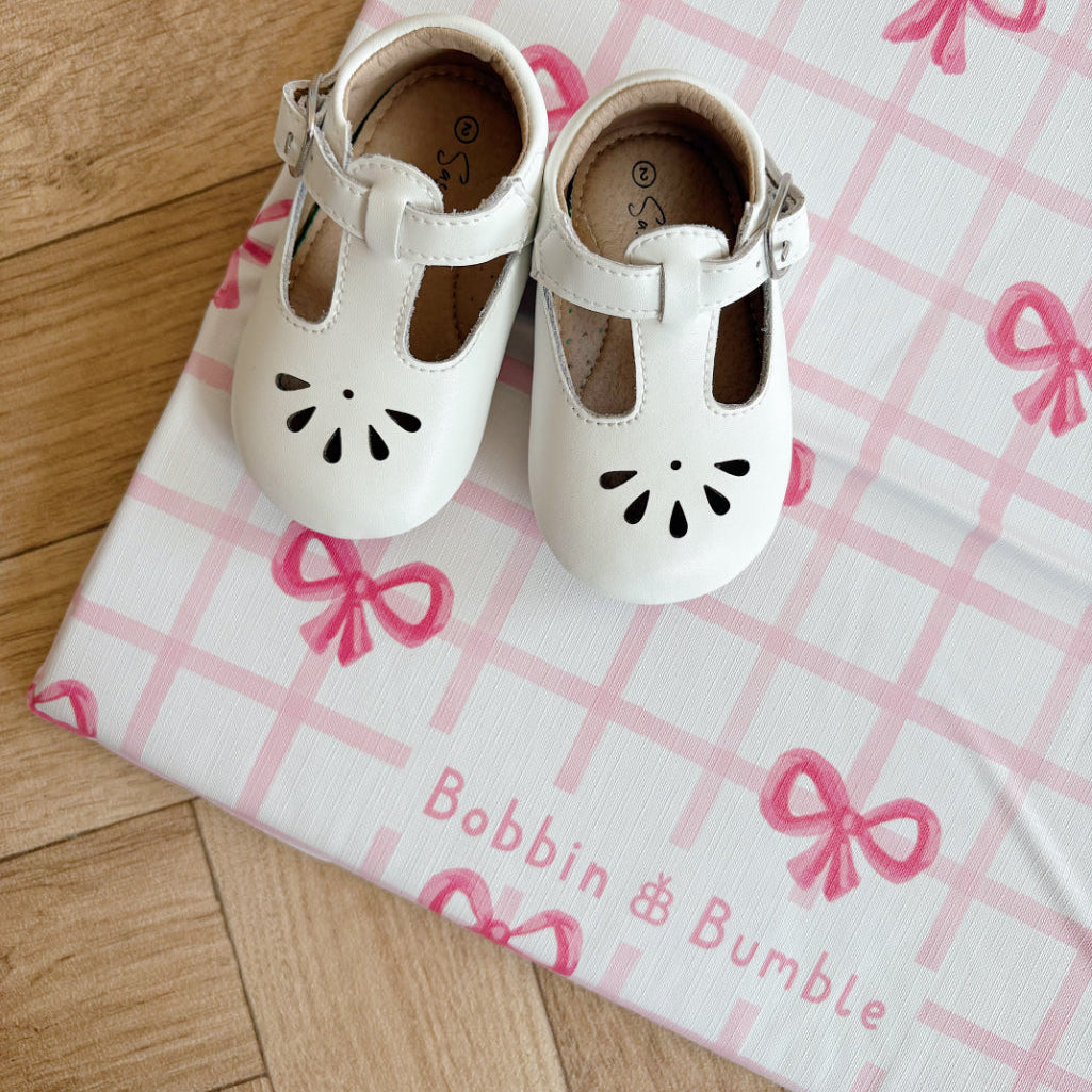 White children's shoes on a pink and white checkered changing mat with bows.