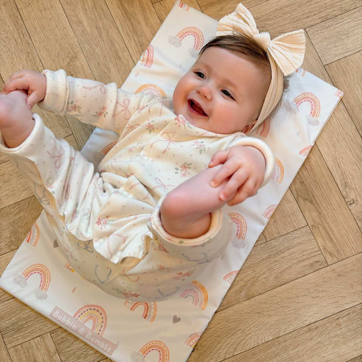 Happy baby wearing a bow headband laying on a rainbow print Bobbin and Bumble travel changing mat on a wooden floor
