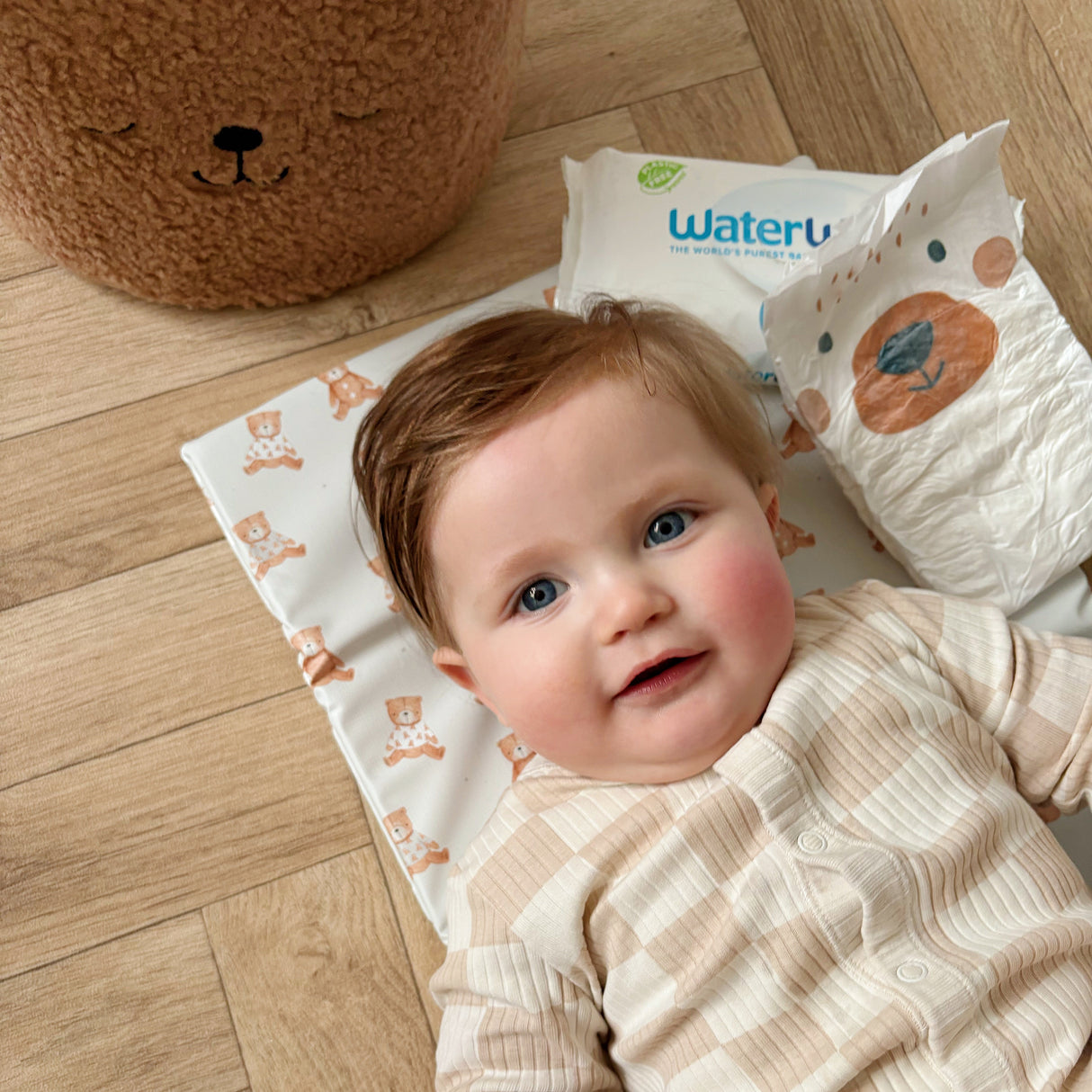 Baby laying on a teddy bear design changing mat with nappy and wipes near by on wooden floor