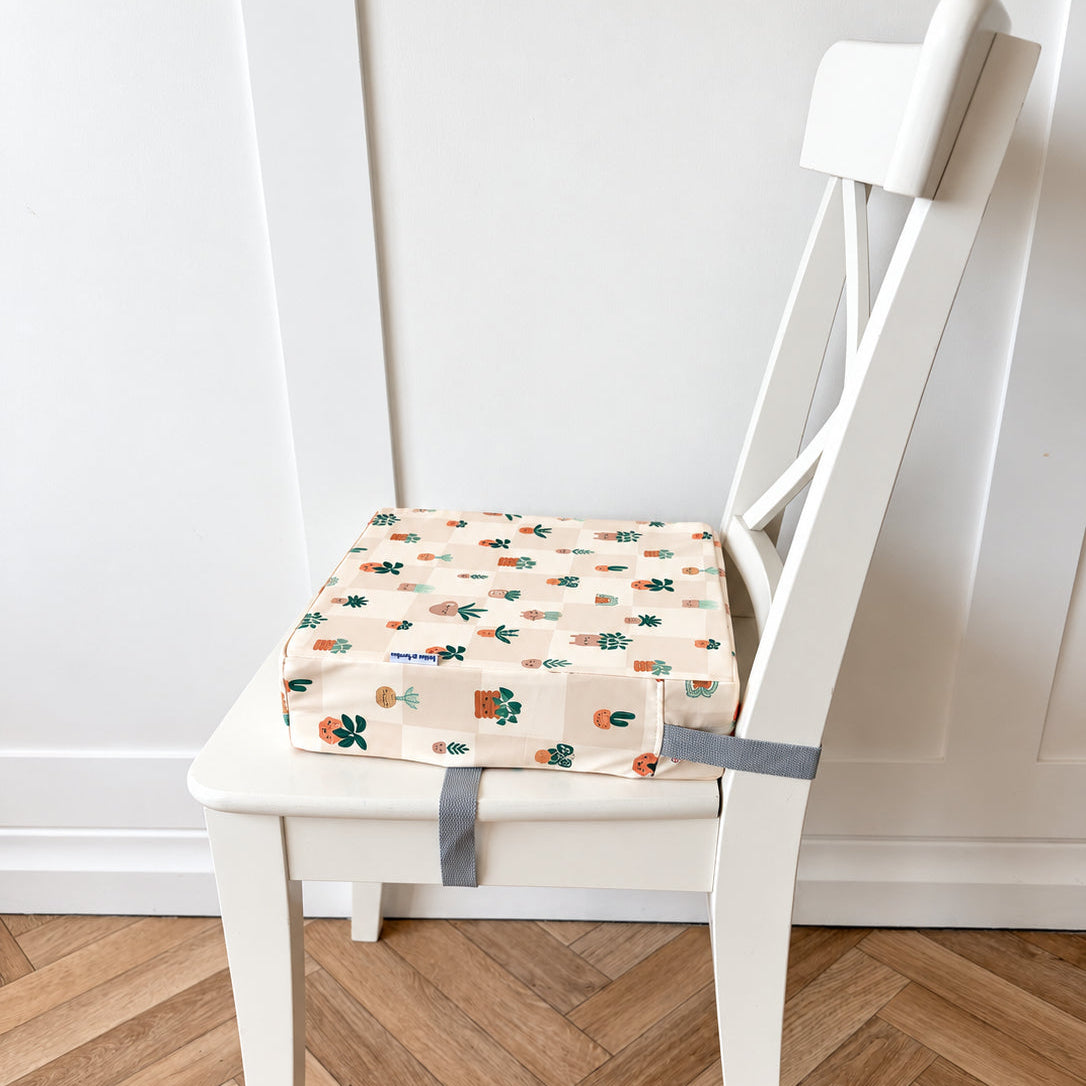 A square fabric chair booster cushion with a colourful plant pattern on a cream background, secured with a grey strap to a white wooden chair on a herringbone wood floor.