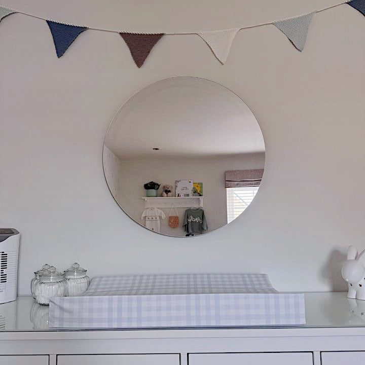 Round mirror on a white wall above a baby changing table with a blue gingham wedge changing mat