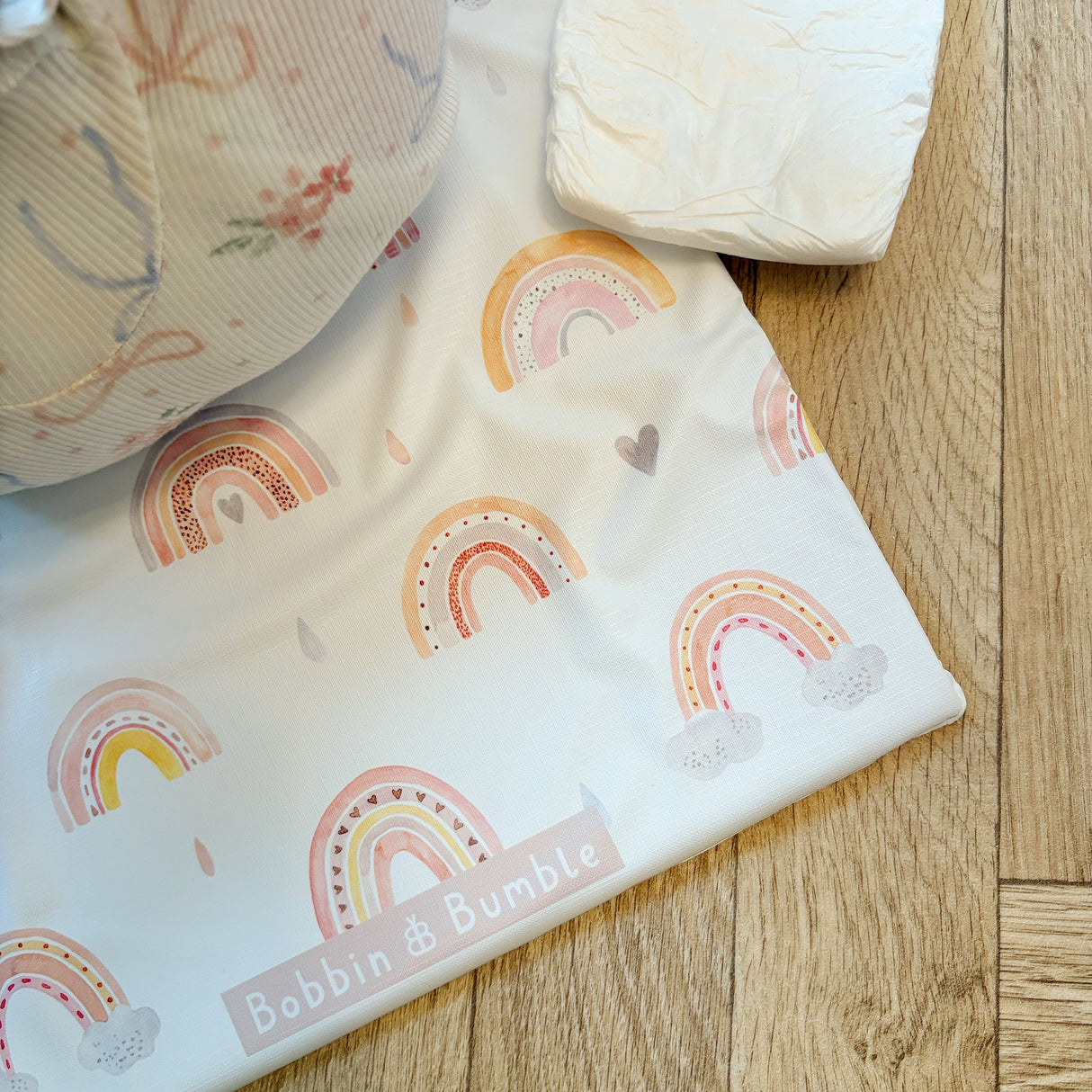 Rainbow print travel changing mat on wooden floor with a nappy close by. Bobbin and Bumble brand visible