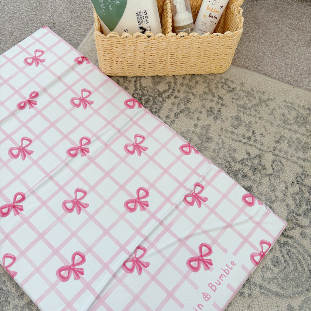 Pink and white checkered baby changing mat with pink bows and 'Bobbin & Bumble' branding, placed on a textured surface.