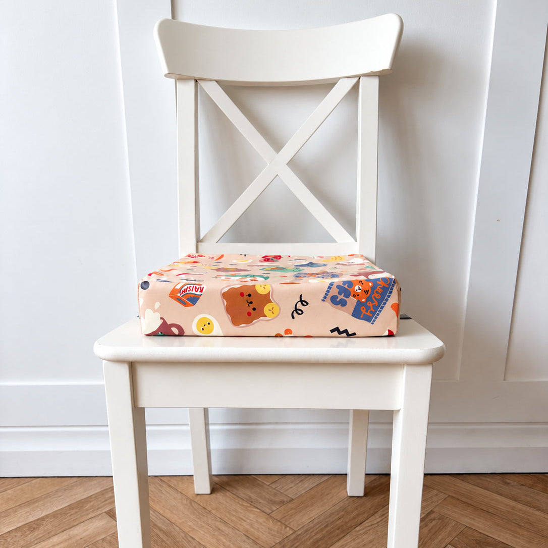 A square booster cushion with a colourful cartoon pattern sits on a white wooden chair in front of a white paneled wall and herringbone wood floor.