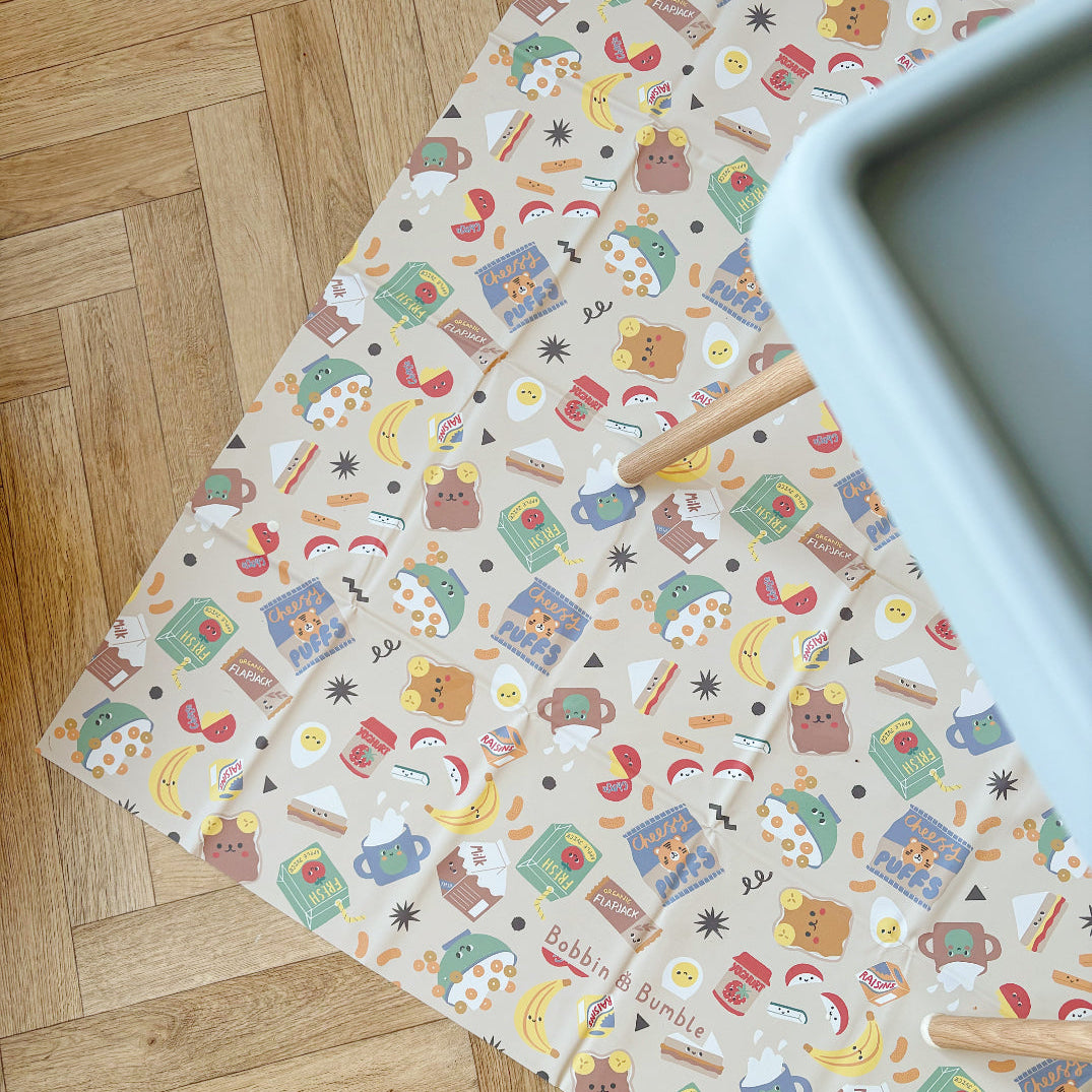 Food-patterned splash mat on a wooden floor with sage green highchair tray partially visible