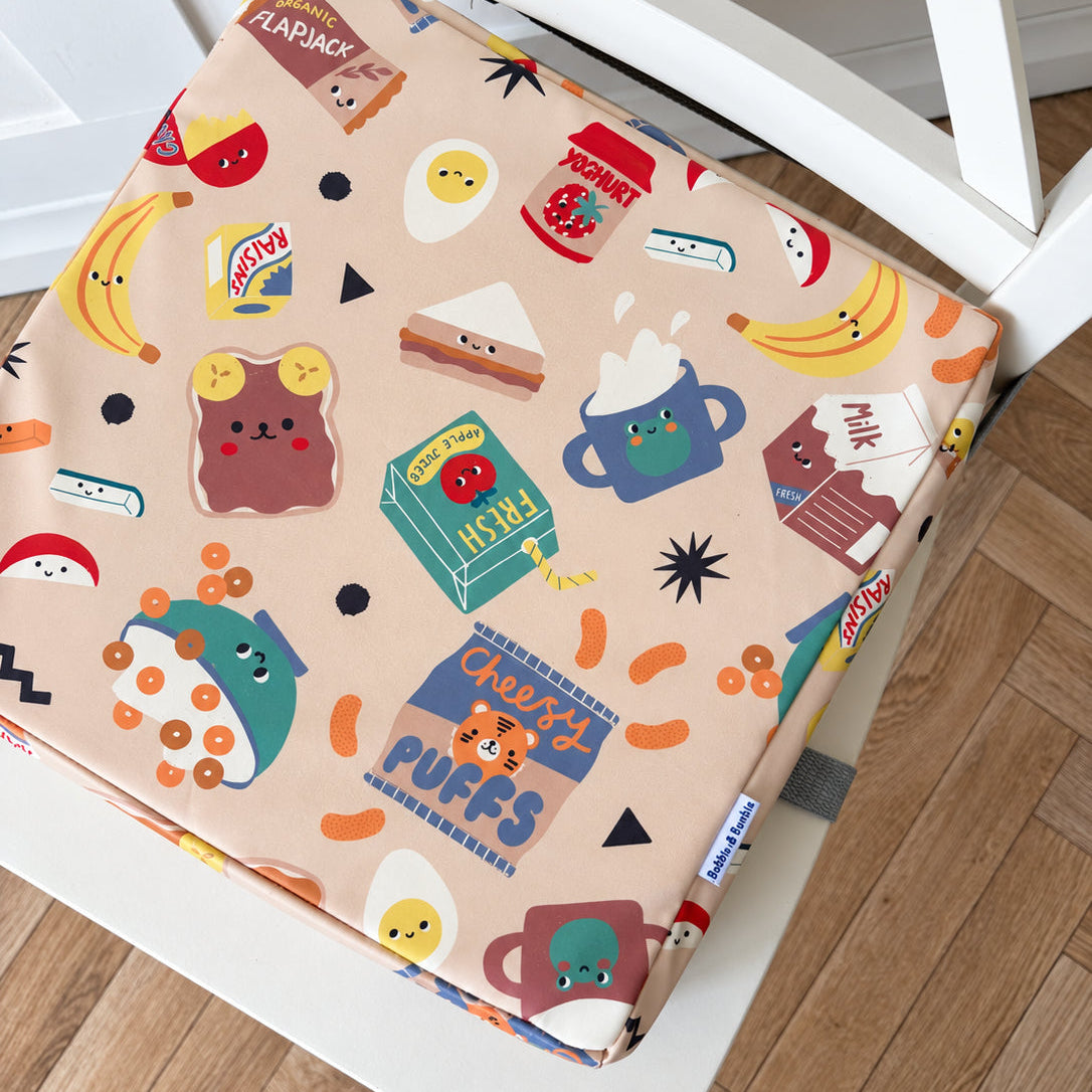 A square fabric booster cushion with a whimsical cartoon food and drink print sits on a white wooden chair on a light wood floor. The print features cute characters and items like bananas, toast, coffee cups, and cereal boxes on a warm beige background.