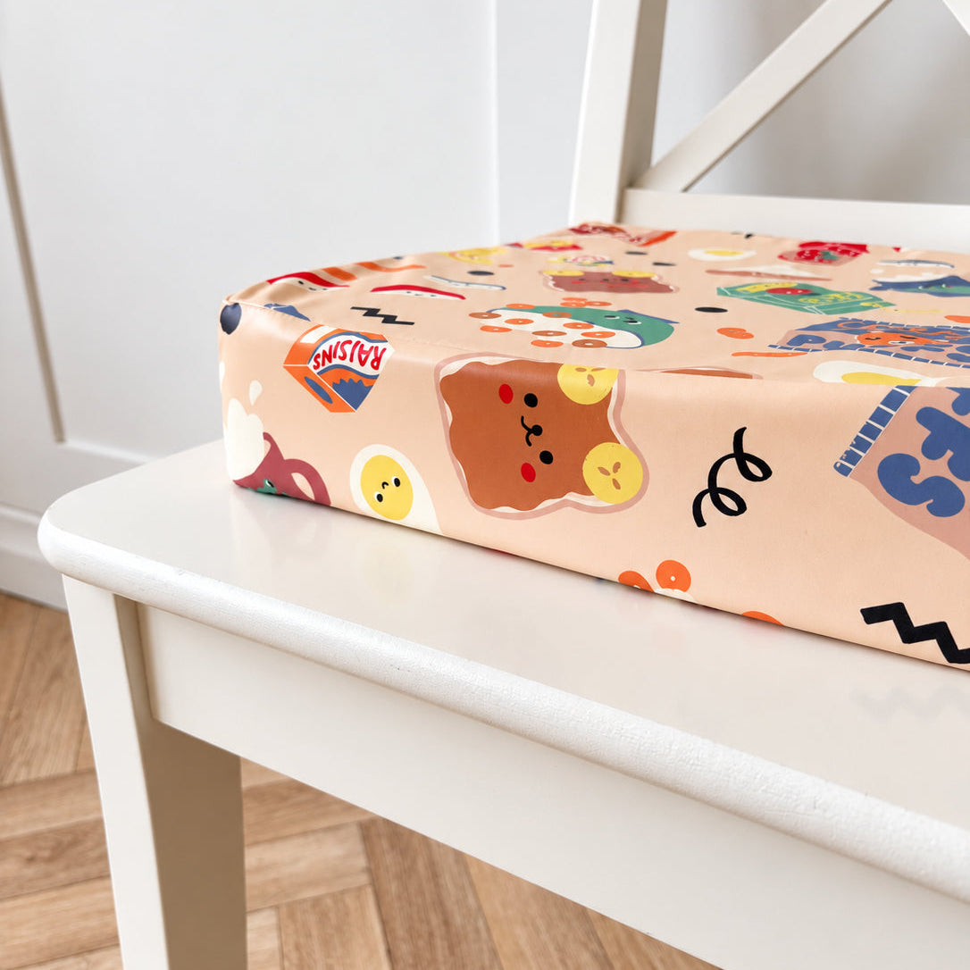 A close-up of a square toddler booster cushion with a colourful cartoon print, resting on a white wooden chair in a bright indoor room with wood flooring.