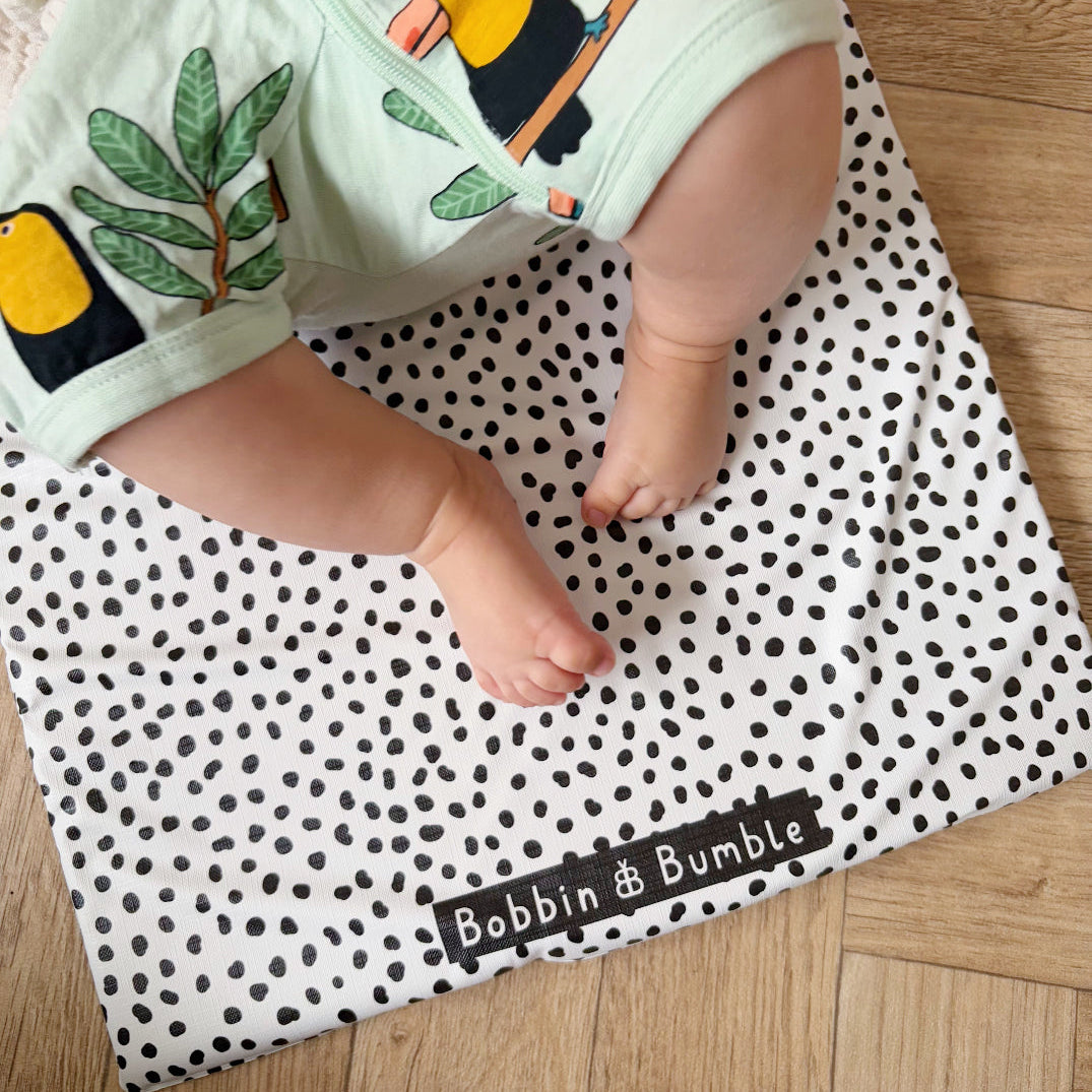 Baby wearing green shorts with toucan design on a polka dot changing mat, brand 'Bobbin & Bumble' visible.