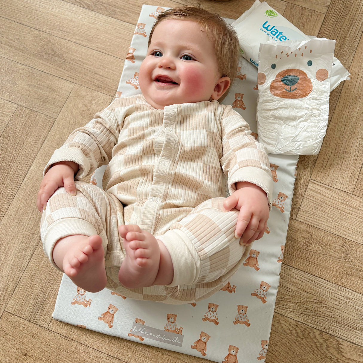 Baby smiling laying on a changing mat with teddy bears design on a wooden floor. Nappy and wipes near by.