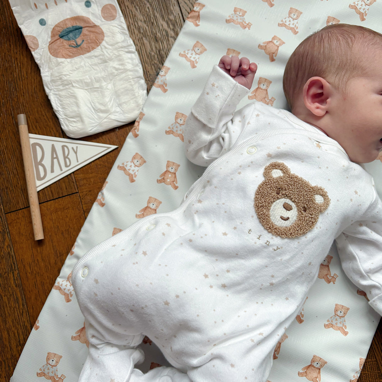 Tiny baby in a teddy bear onesie laying on a teddy bear changing mat with a nappy and a pennant flag saying 'baby' close by on a wooden floor.