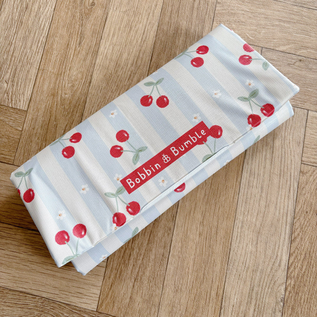Folded baby changing mat with cherry pattern and Bobbin & Bumble logo on a wooden floor.