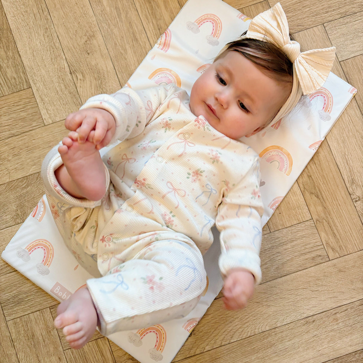 Happy baby wearing a bow headband laying on a rainbow print Bobbin and Bumble travel changing mat on a wooden floor