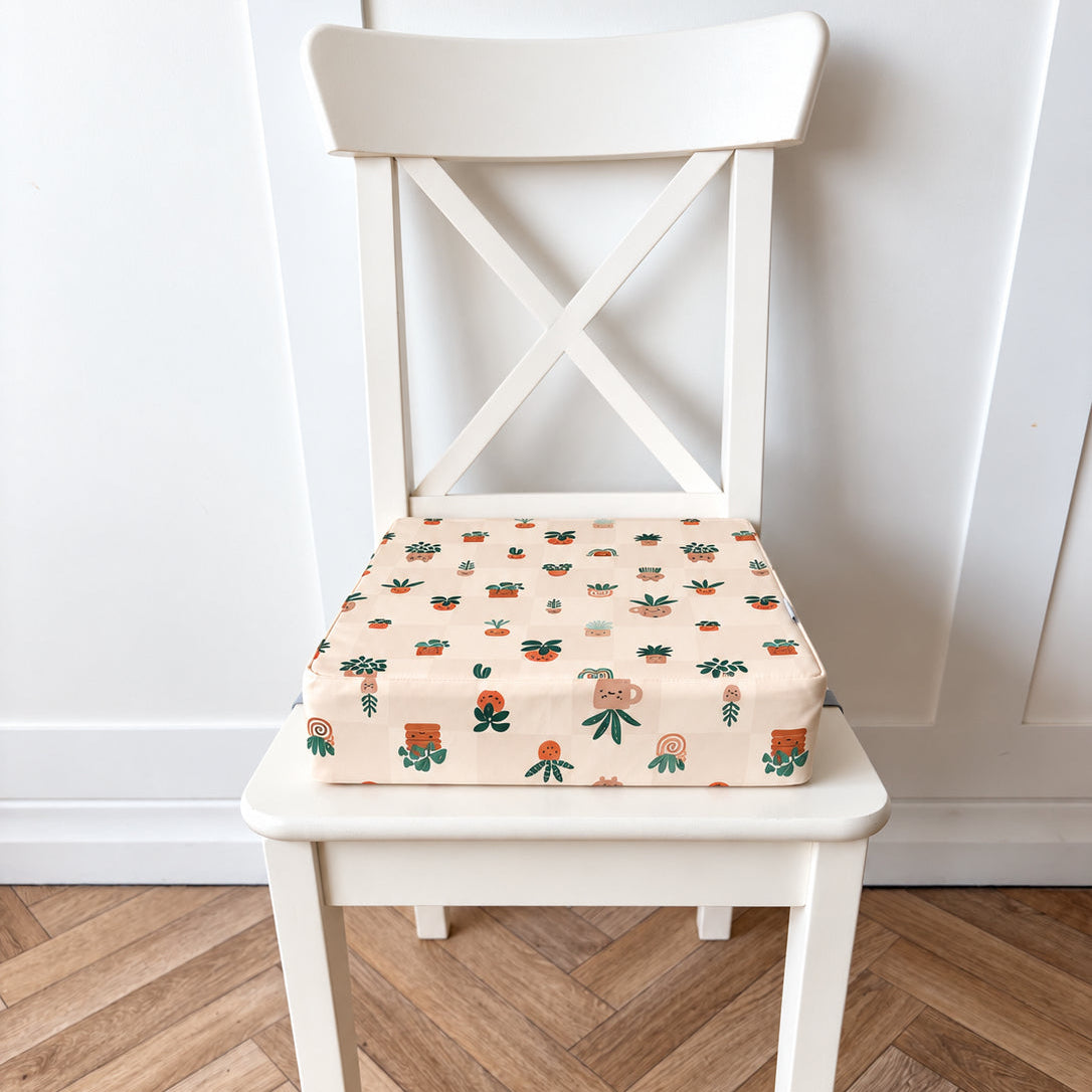 A square booster seat cushion with a colourful potted plant pattern on a cream background, shown on a white wooden chair in a room with a light wood herringbone floor and white paneled wall.