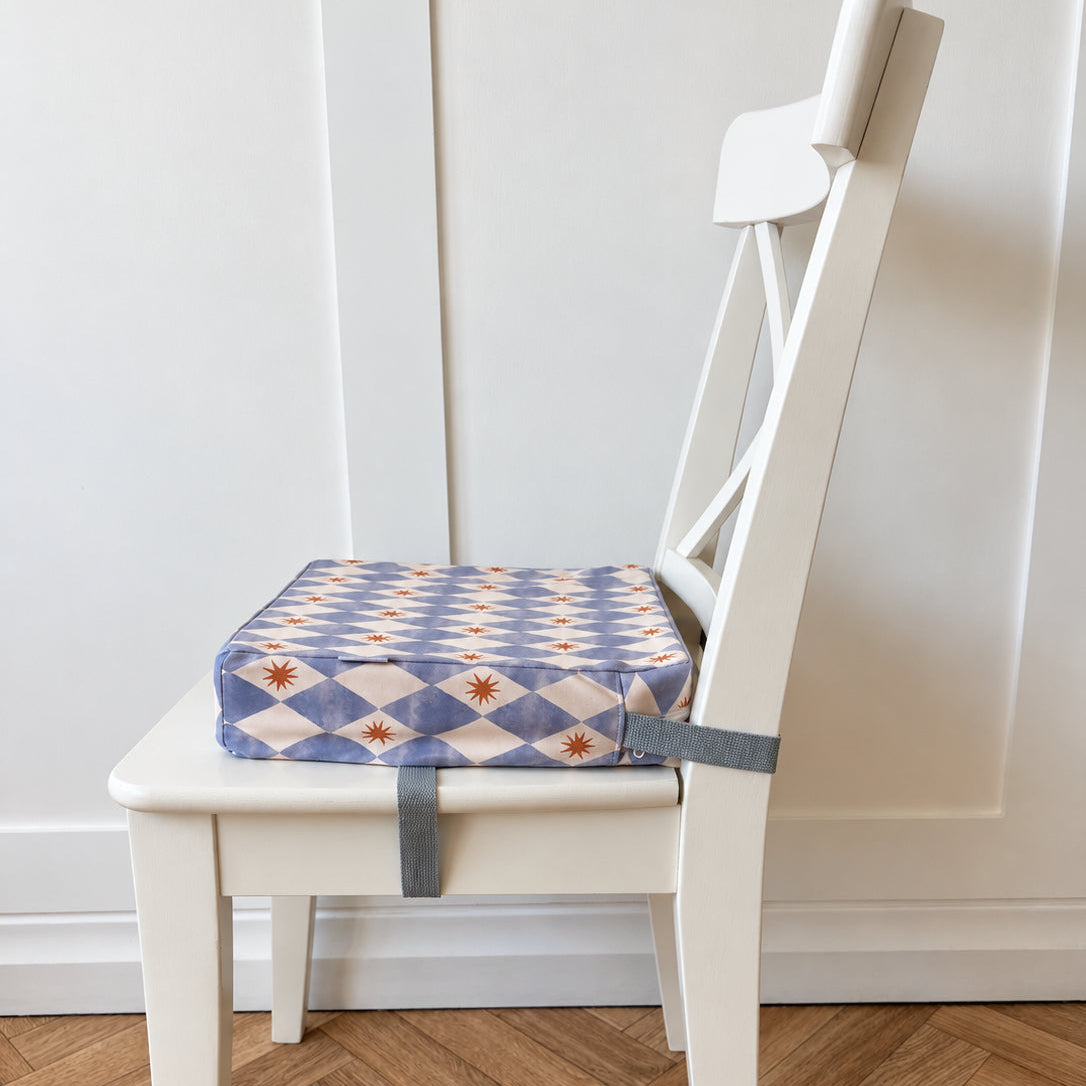 A square fabric booster chair cushion with a blue, white, and yellow diamond pattern, strapped to the seat of a white wooden dining chair on a light wood herringbone floor.